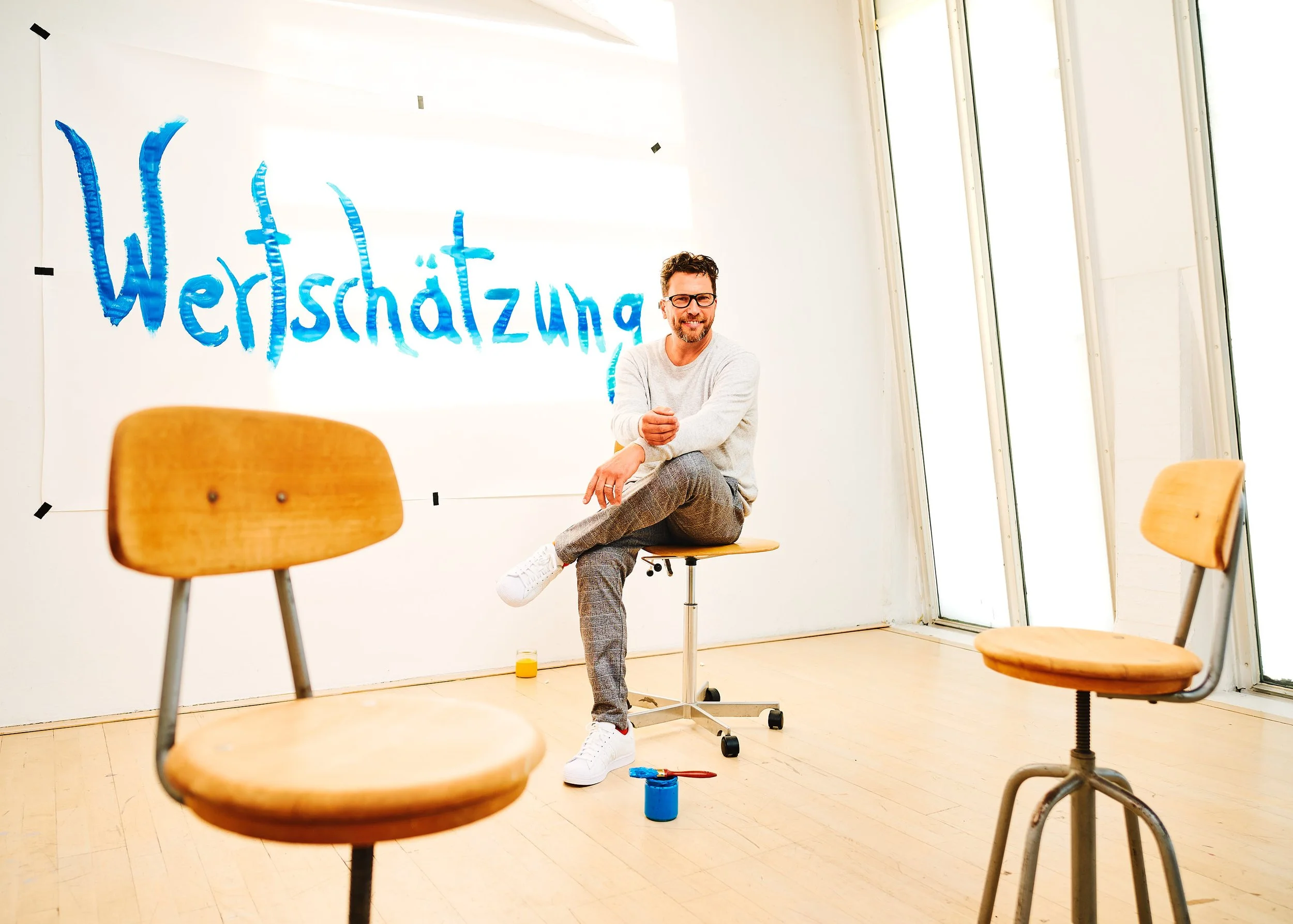 Der Coach Julian Böhme sitzt auf einem Stuhl in einem Raum mit großen Fenstern. An der Wand steht das Wort „Wertschätzung“ in blauer Farbe. Es gibt zwei leere Stühle im Raum und Farbeimer auf dem Boden.