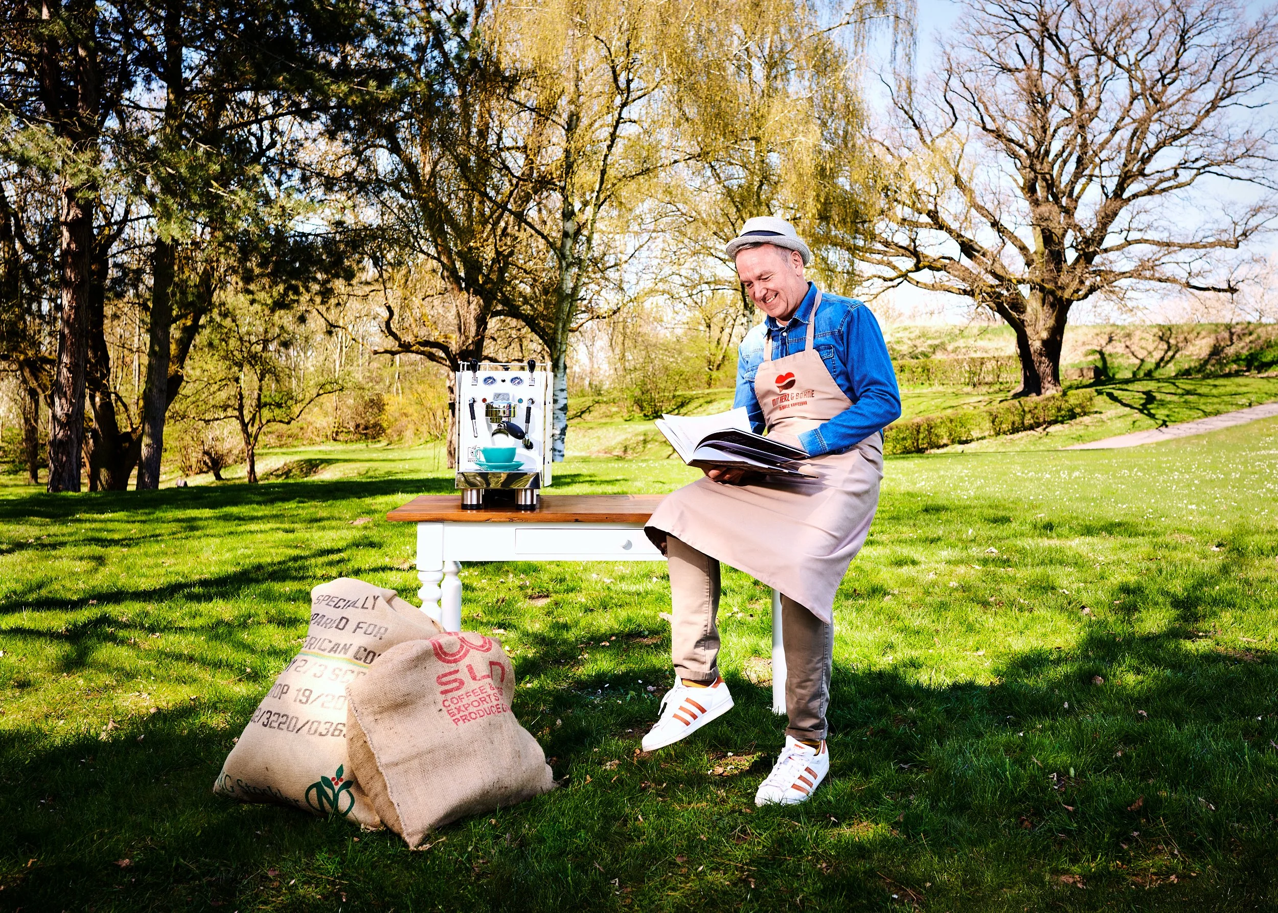 Götz Ohlendorf sitzt im freien auf einem Stuhl und liest ein Buch. Er trägt eine Schürze und weiße Turnschuhe. Auf einem Tisch steht eine Espressomaschine. Auf dem Boden liegen zwei große Säcke mit Kaffeebohnen, umgeben von Bäumen und grünem Gras.
