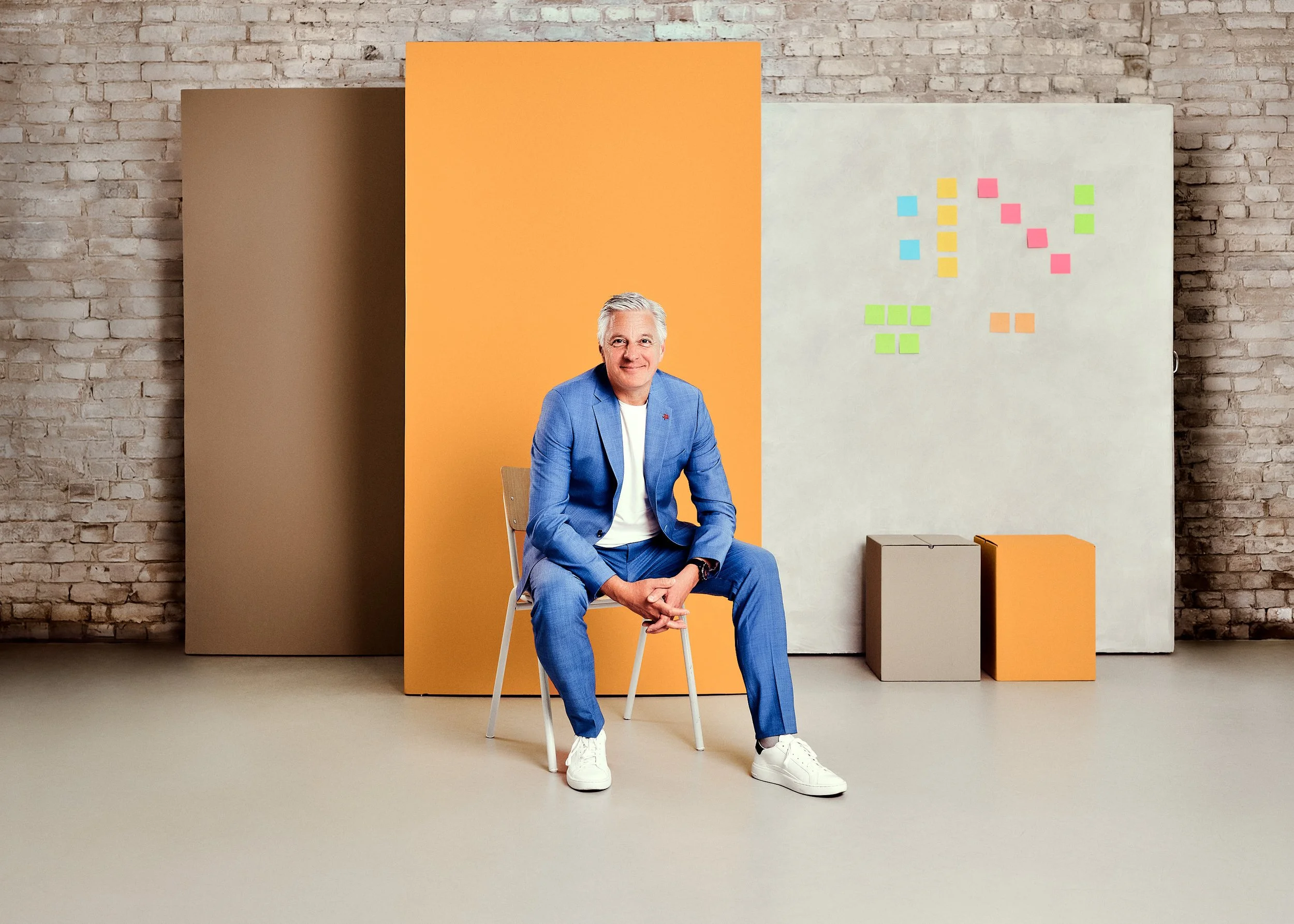 Ein älterer Mann in blauer Anzug sitzt auf einem Stuhl vor bunten Hintergründen mit Post-its und rechteckigen Objekten, mit einer Brickwände im Hintergrund.