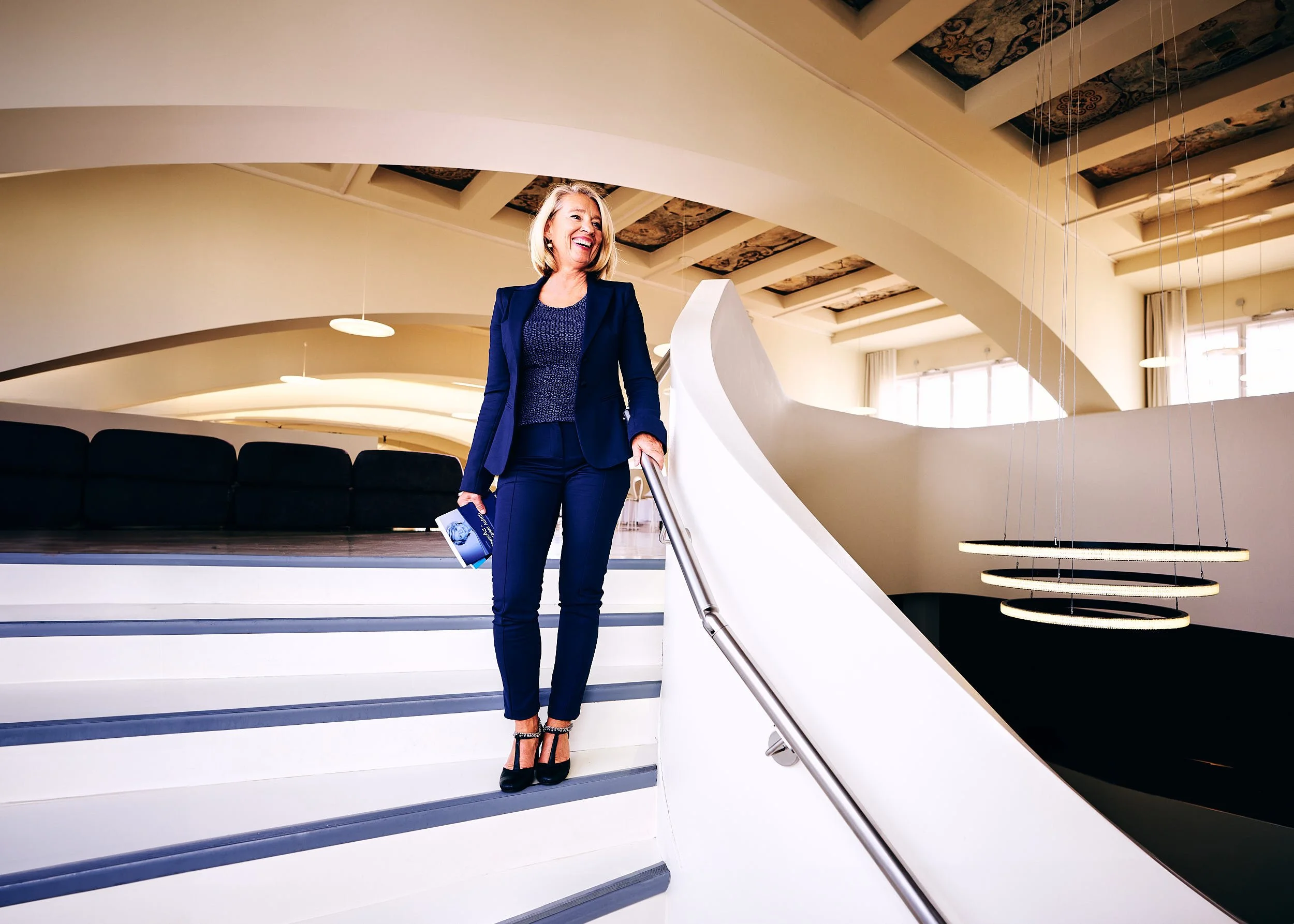 Eine fröhliche Frau in einem blauen Hosenanzug steht auf einer weißen Treppe im modernen, hellen Innenraum und hält ein Magazin in der Hand.