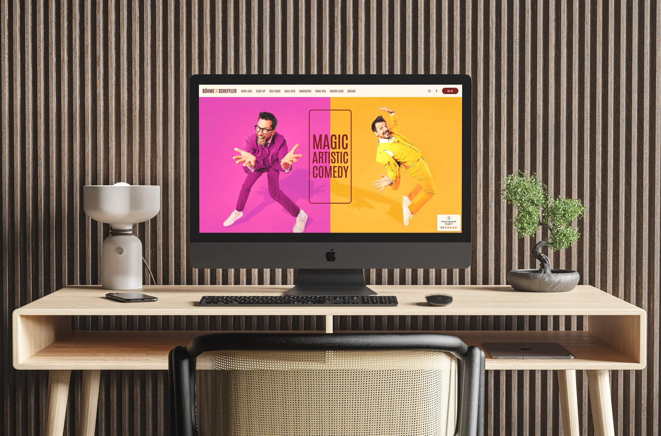 Ein Computerbildschirm mit buntem Werbeplakat für Comedy-Shows, flachem Holzschreibtisch, weißer Vase mit Pflanze, schwarzer Maus und Laptop, vor einer Wand mit vertikalen Holzpaneelen.