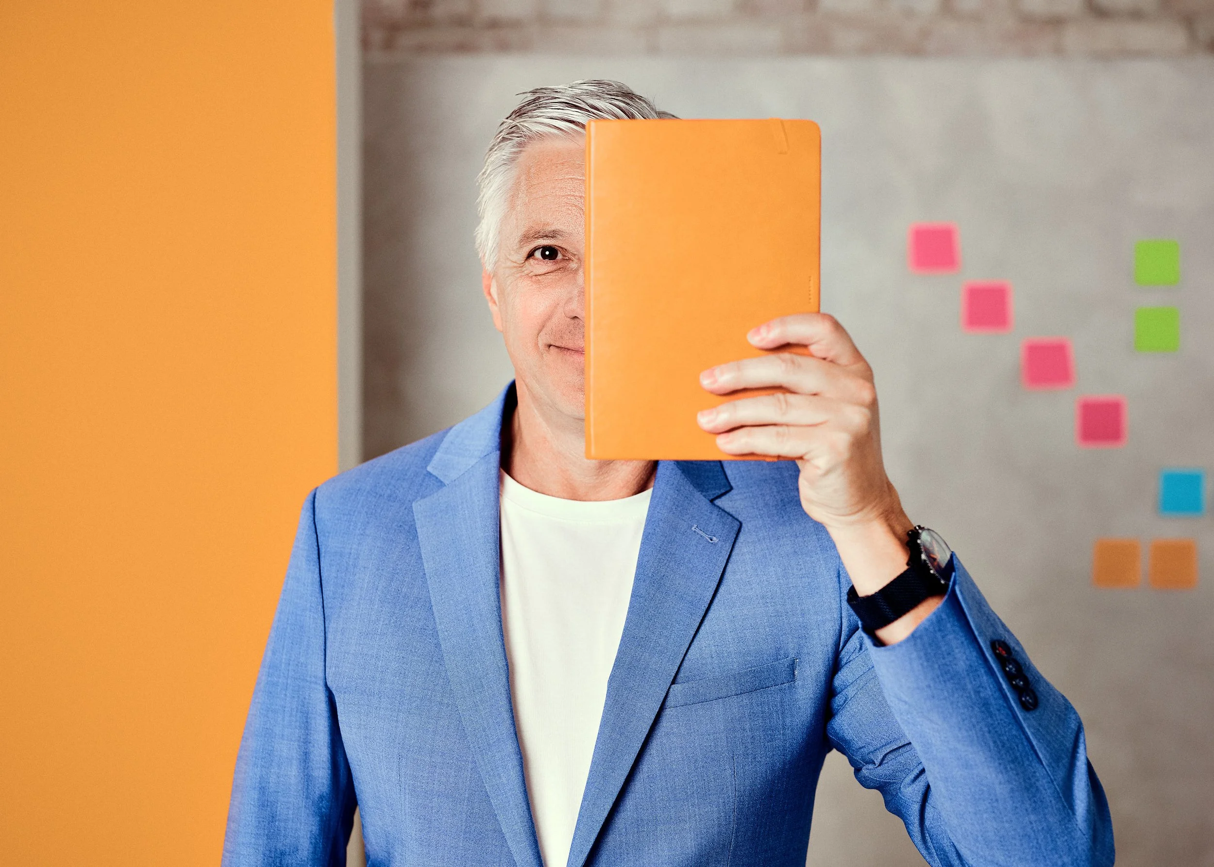 Ein Mann in einem blauen Anzug hält einen orangefarbenen Ordner vor sein Gesicht, sodass nur ein Auge und ein Teil seines Gesichts sichtbar sind, im Hintergrund sind bunte Post-its an einer grauen Wand.