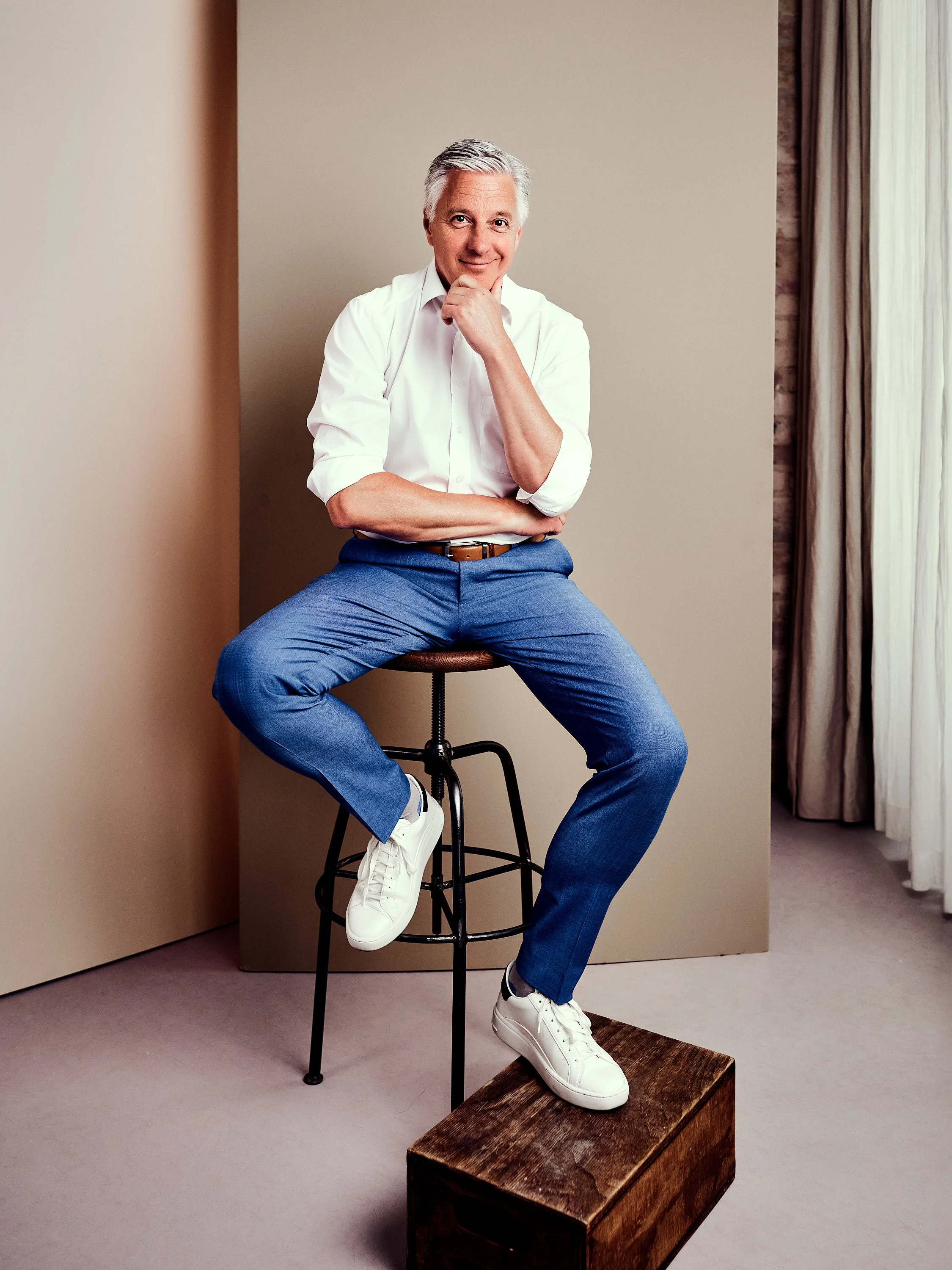Ein älterer Mann sitzt auf einem Barhocker, seine Beine sind auf einer Holztruhe platziert. Er trägt ein weißes Hemd, blaue Hosen und weiße Sneakers. Der Mann hat graues Haar, lächelt und hat die rechte Hand am Kinn, während er in die Kamera schaut. Der Hintergrund ist einfach mit einer neutralen Wand und Vorhängen auf der rechten Seite.