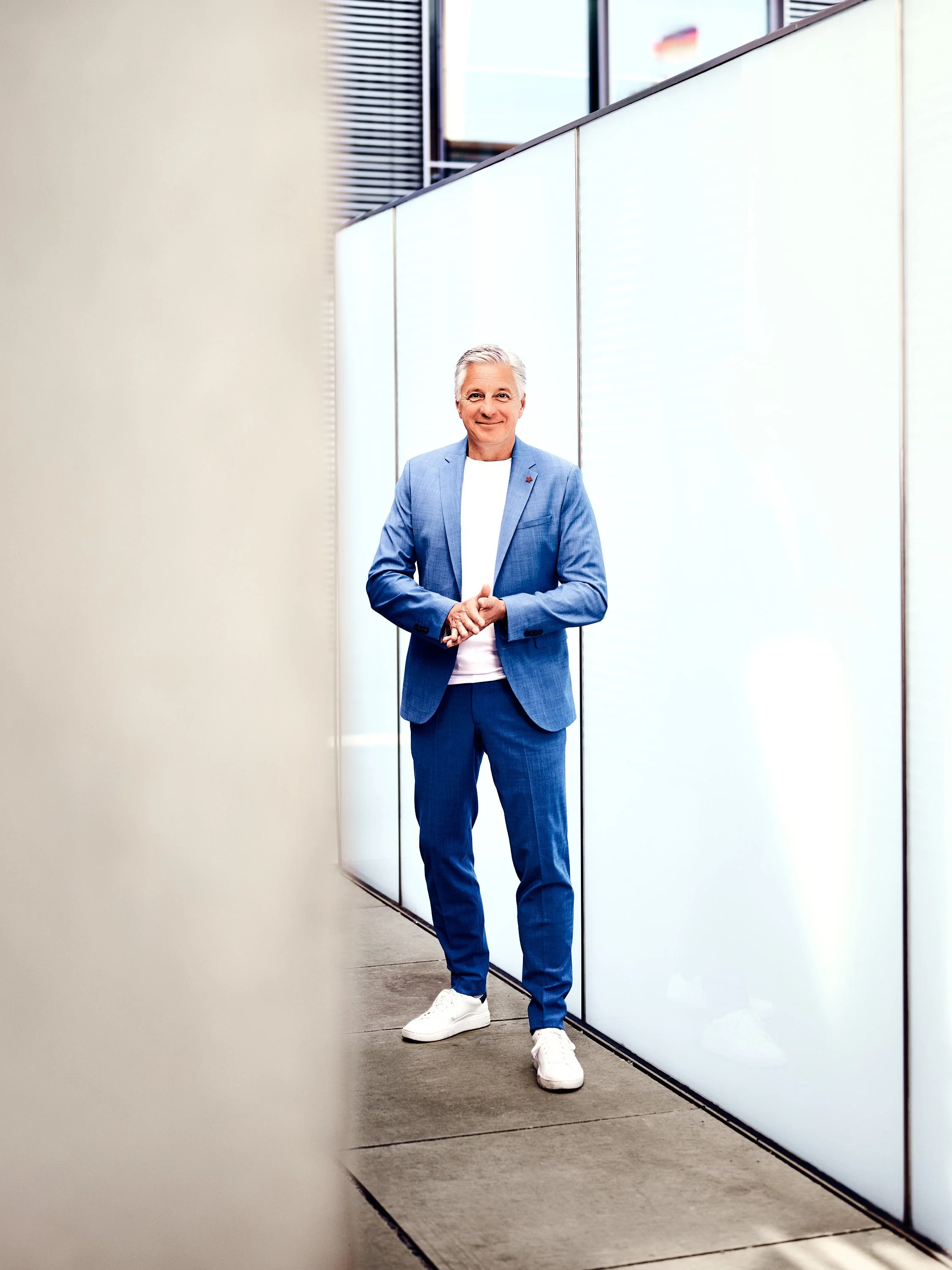 Ein Mann in einem blauen Anzug, weißen Turnschuhen und einem weißen T-Shirt steht vor einer beleuchteten Wand in einem modernen Gebäude und lächelt.