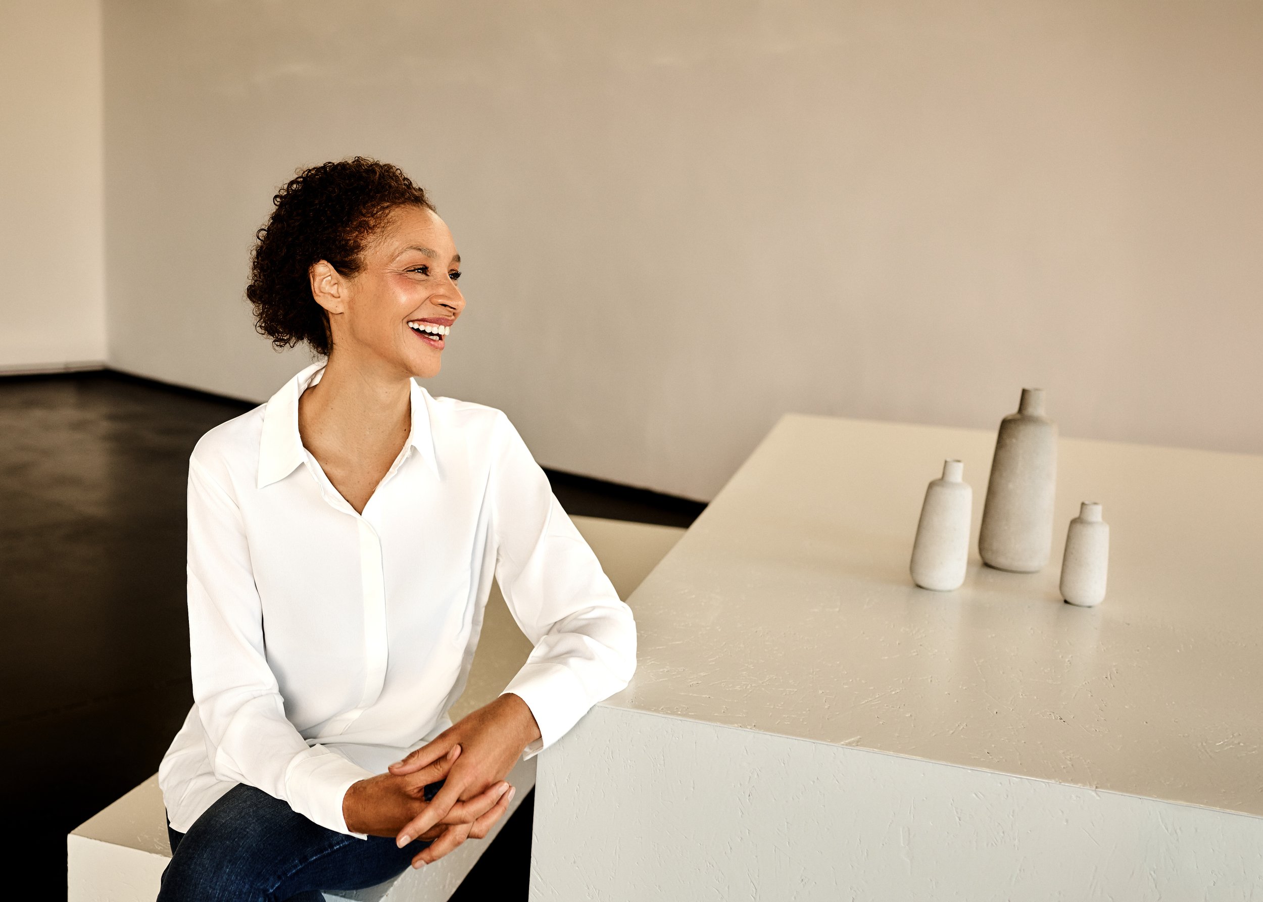 Eine lachende Frau in weißem Hemd sitzt an einer weißen Theke in einem modernen Raum mit minimalistischer Dekoration und vier grauen Keramikvasen.