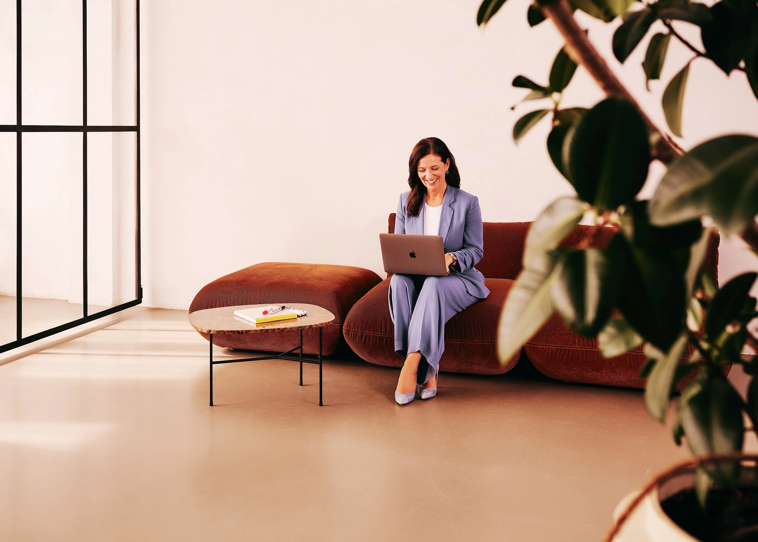 Eine lachende Frau sitzt auf einem roten Sofa, trägt einen grauen Anzug, benutzt einen Laptop und hat auf einem Couchtisch Stifte und eine Notiz. Es ist ein modernes, helles Raum mit weißen Wänden und einer Pflanze im Vordergrund.