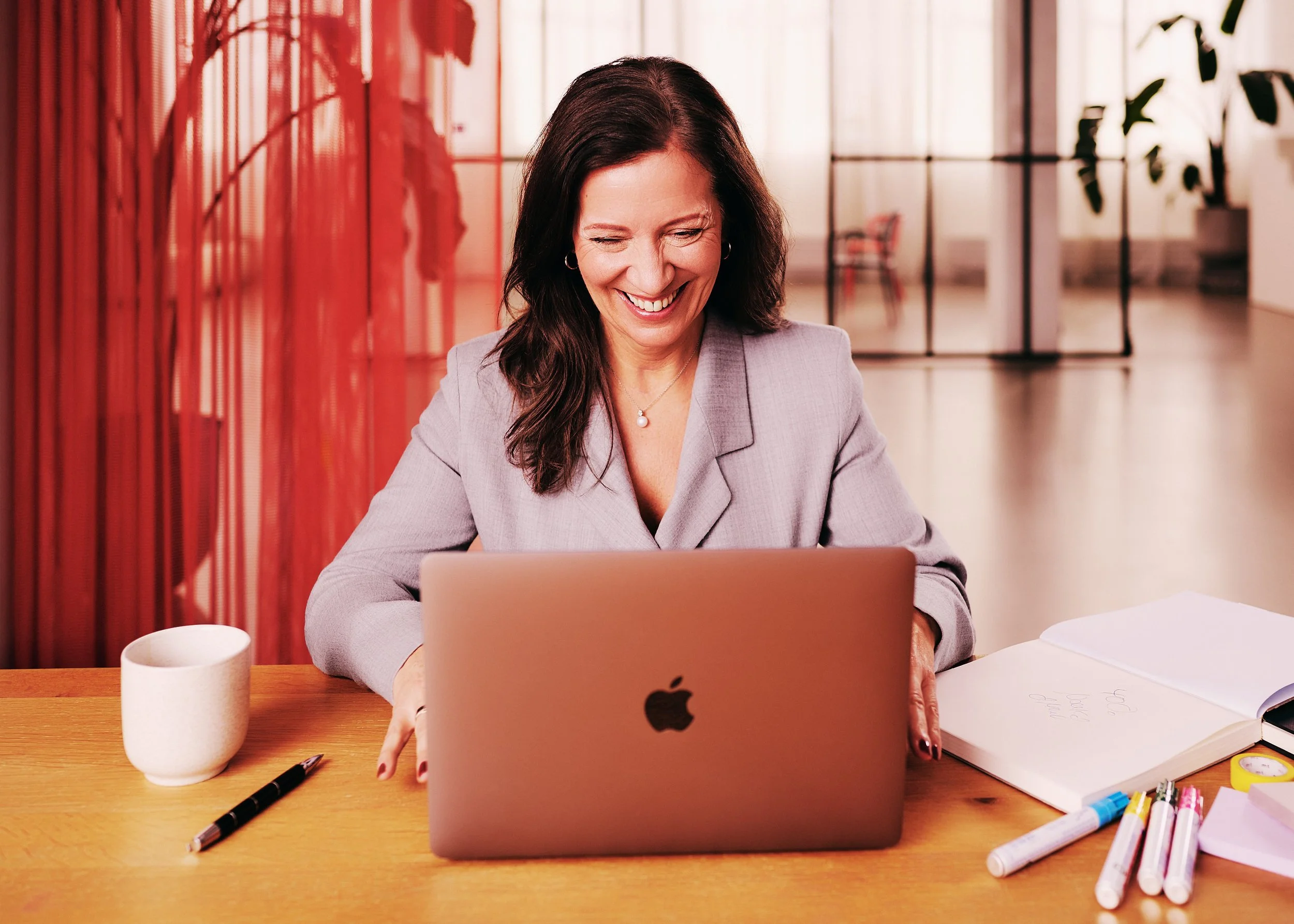 Eine lachende Frau arbeitet an einem Laptop in einem modernen Raum mit großen Fenstern und Pflanzen im Hintergrund, umgeben von Notizzetteln, Markern und einer Kaffeetasse auf einem Holztisch.