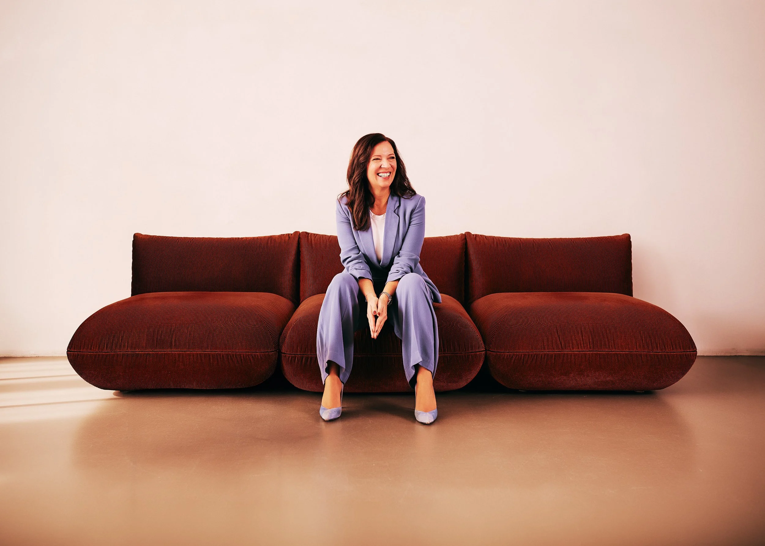 Eine lachende Frau in einem lilafarbenen Business-Outfit sitzt auf einer roten Couch vor einer weißen Wand.