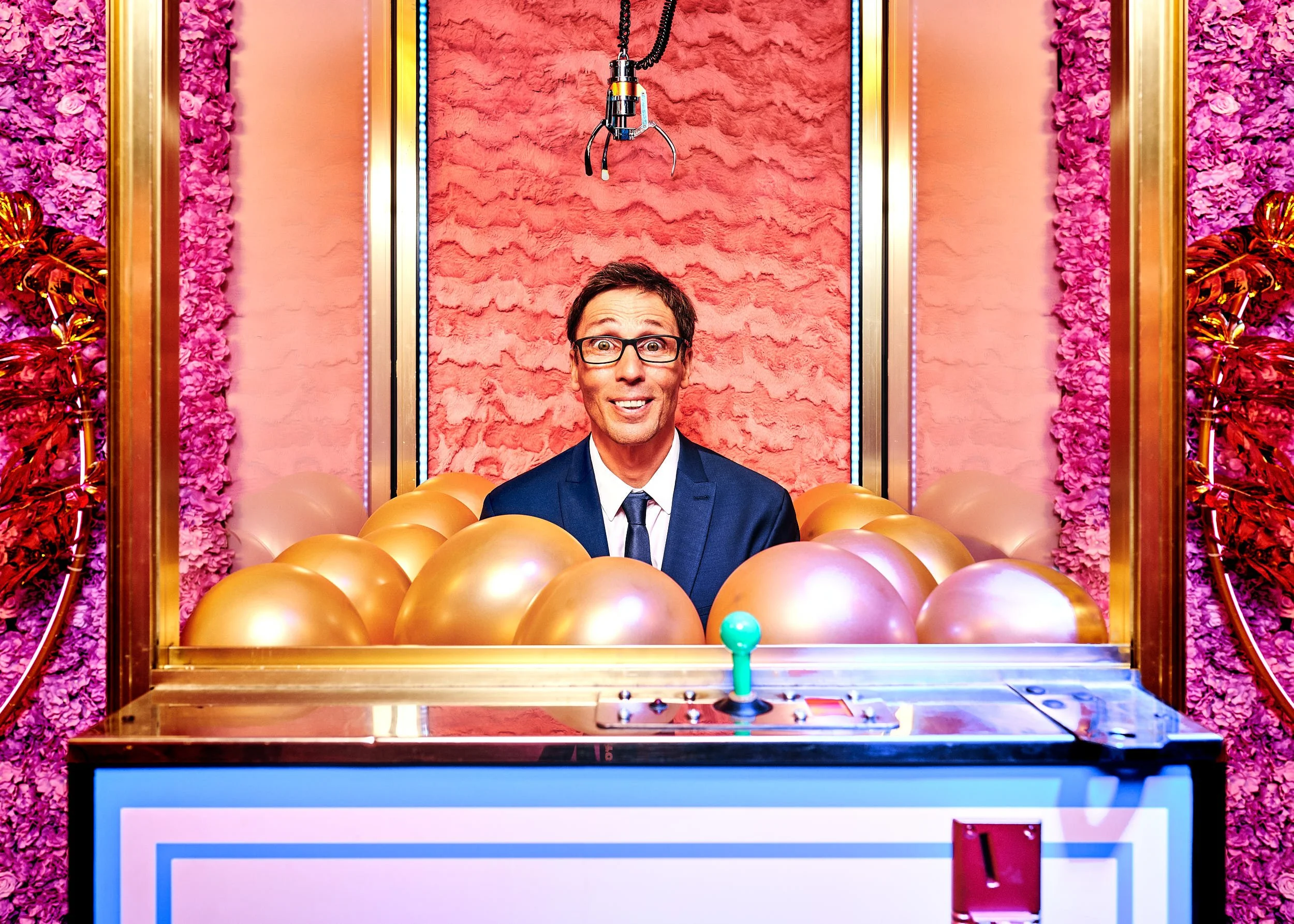 Ein Mann in Anzug und Brille sitzt vor einer Rummel- oder Spielhalle-Kulisse mit rosa und goldfarbenen Dekorationen und Pompoms, ein Claw Crane Spiel ist vorne sichtbar, es gibt einen Schläger und eine Kugel im Spiel.