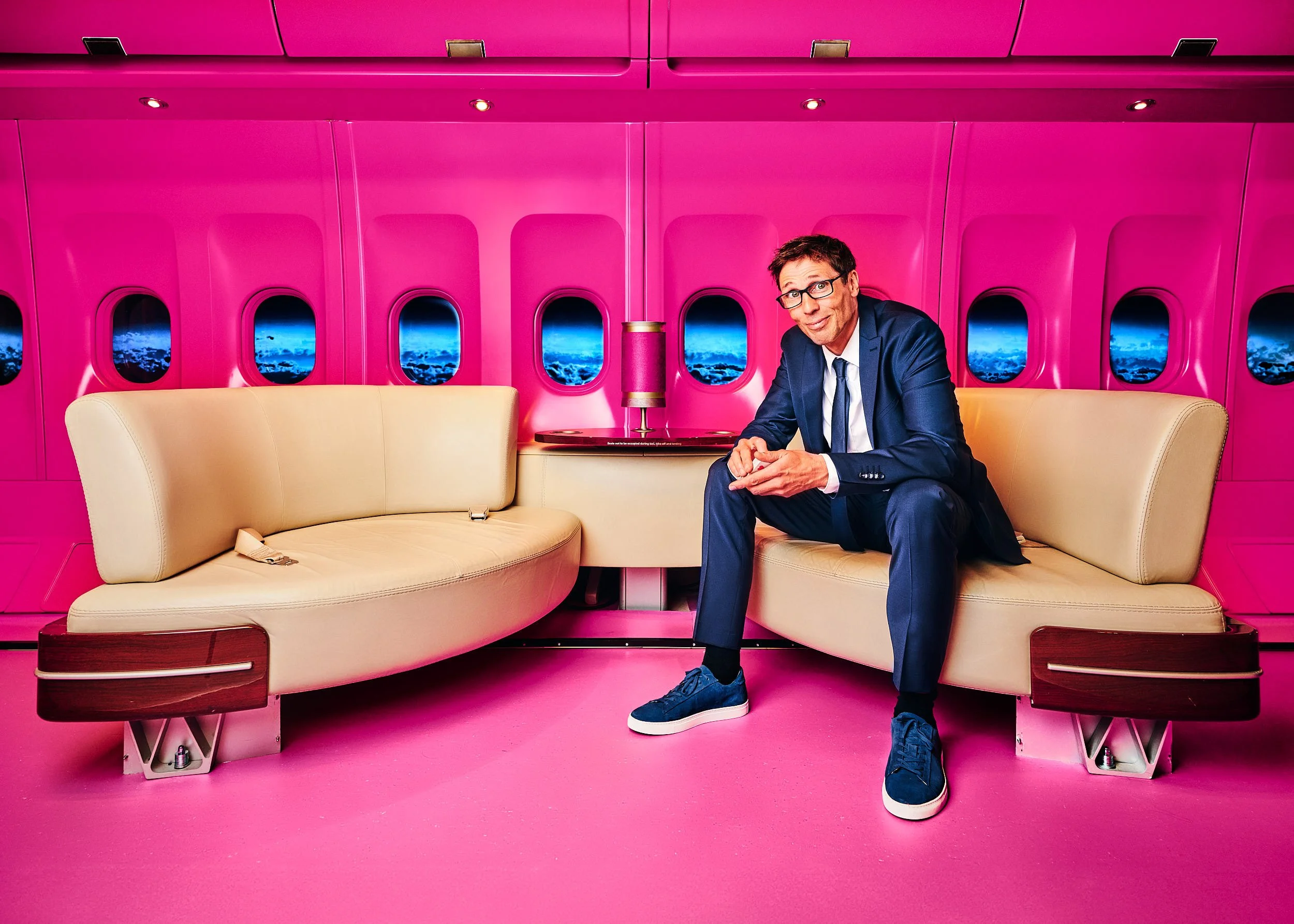 Ein Mann in dunklem Anzug sitzt in einem pinkfarbenen Raum mit Blick auf die Erde aus einem Flugzeugfenster.