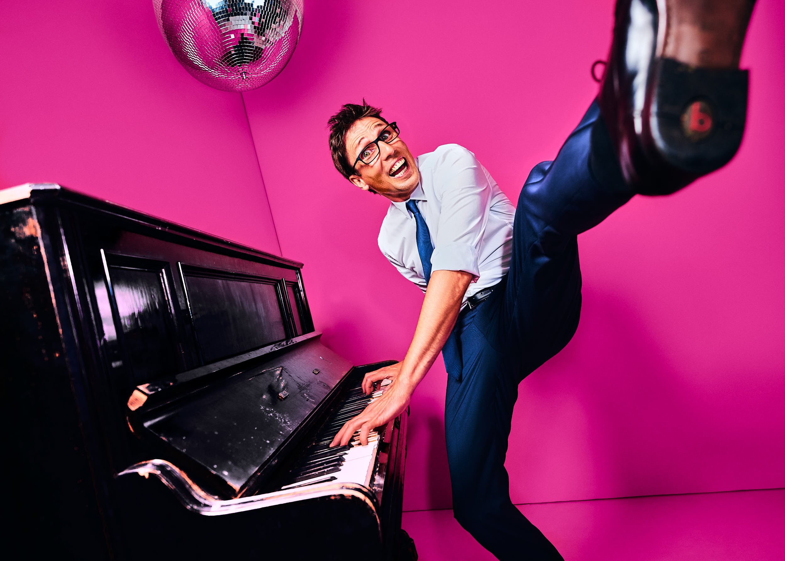 Mann in Anzug und Krawatte spielt auf einem schwarzen Klavier vor pinkem Hintergrund, mit einer Disco-Kugel über ihm.