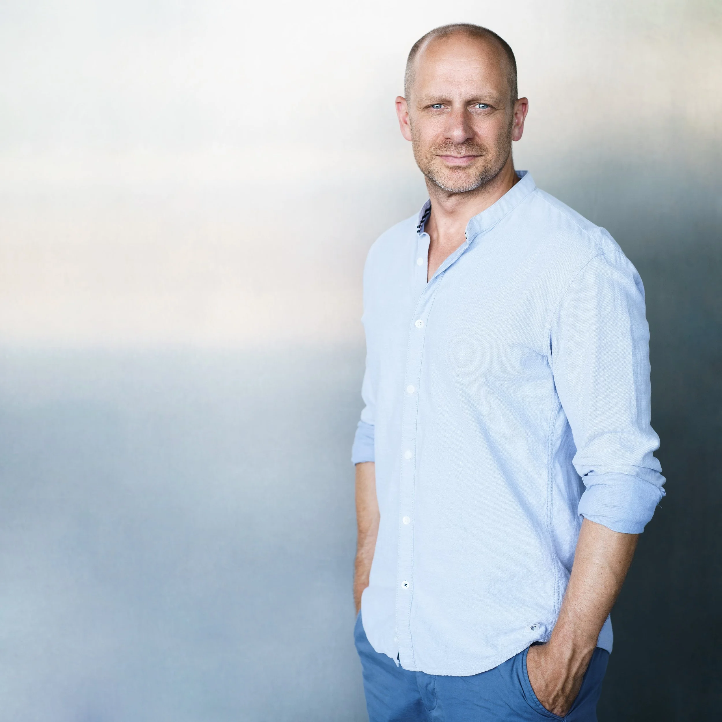 Ein Mann trägt ein hellblaues Hemd und blaue Jeans, steht vor hellem Hintergrund und lächelt leicht.