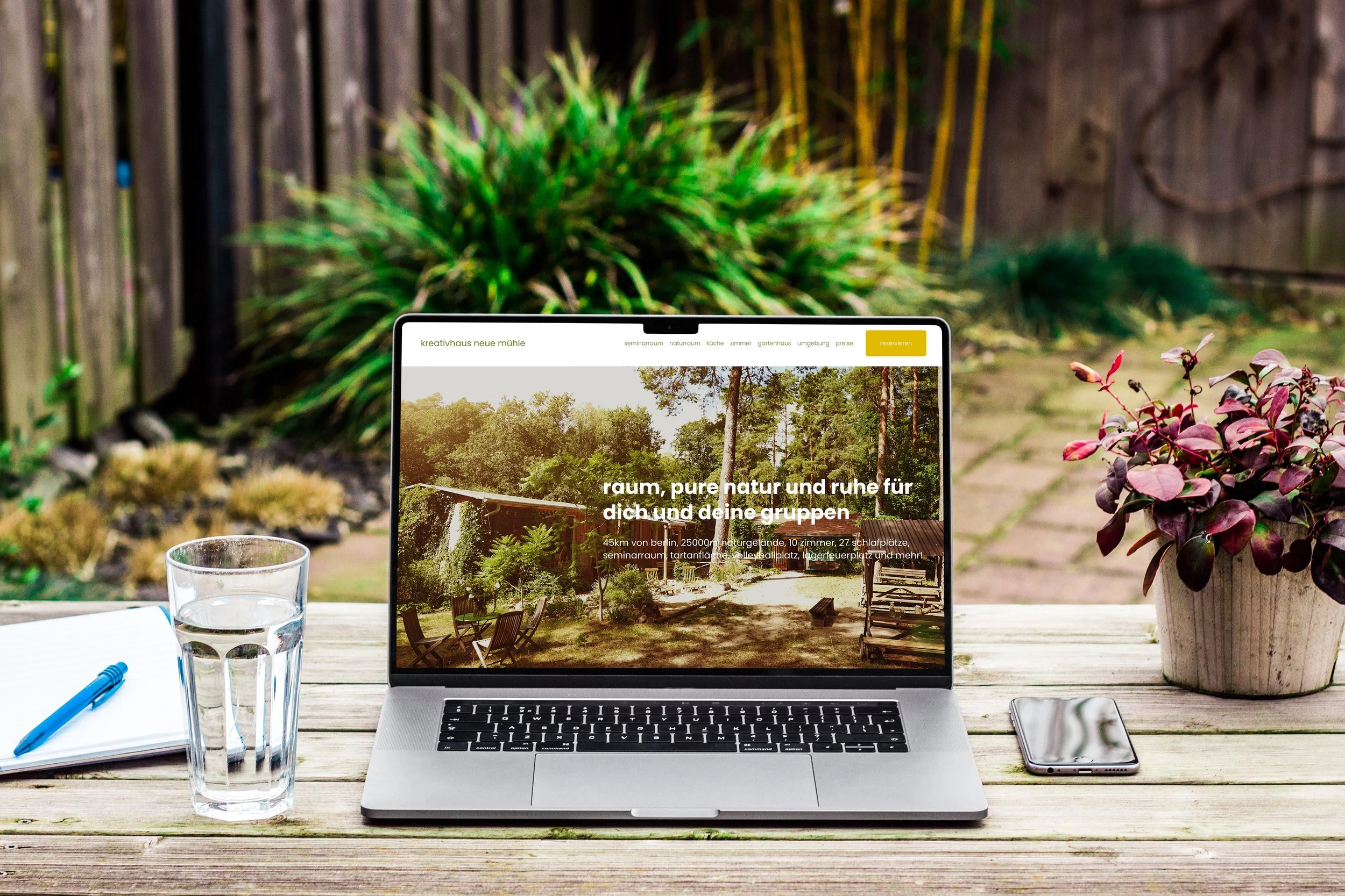 Laptop auf einem Holz Tisch mit Wasser, Notizbuch, Stift, Smartphone, Pflanzen im Garten, Bildschirm zeigt eine Webseite über Naturunterkünfte.