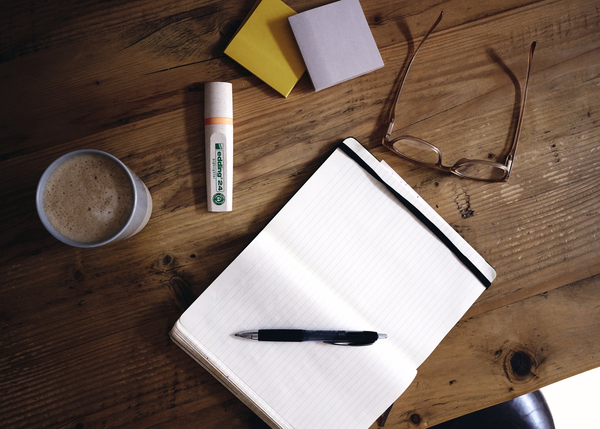 Auf einem Holzschreibtisch liegen eine Tasse Kaffee, ein Whiteboard-Marker, zwei Notizbücher, eine Brille, eine Spiralnotizbuch und ein Kugelschreiber.