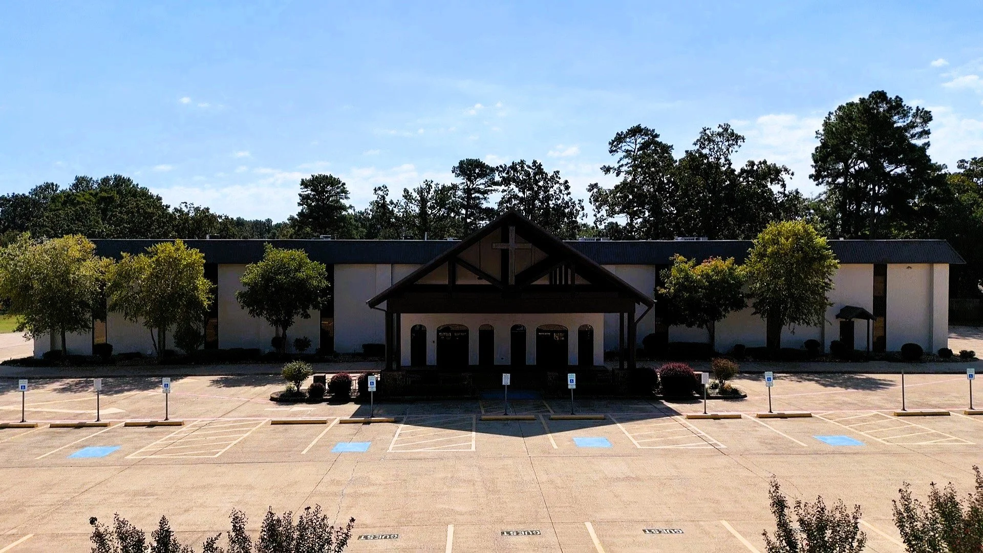 A building with a prominent cross over its entrance, Landmark Pentecostal Church in Texarkana TX, surrounded by trees, with a parking lot in front that has designated handicap parking spots.
