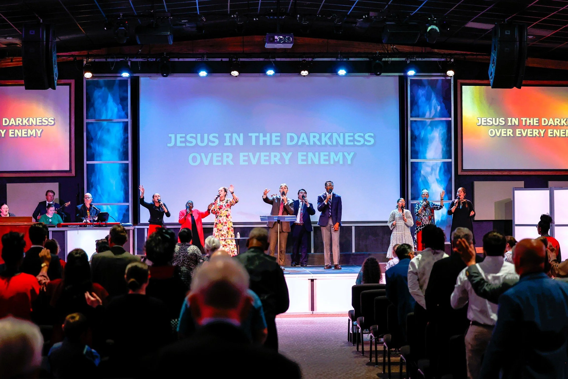 A church service at Landmark Pentecostal Church in Texarkana TX. Large screens behind display the lyrics 'JESUS IN THE DARKNESS OVER EVERY ENEMY.' The audience is visible, seated and participating in the service.