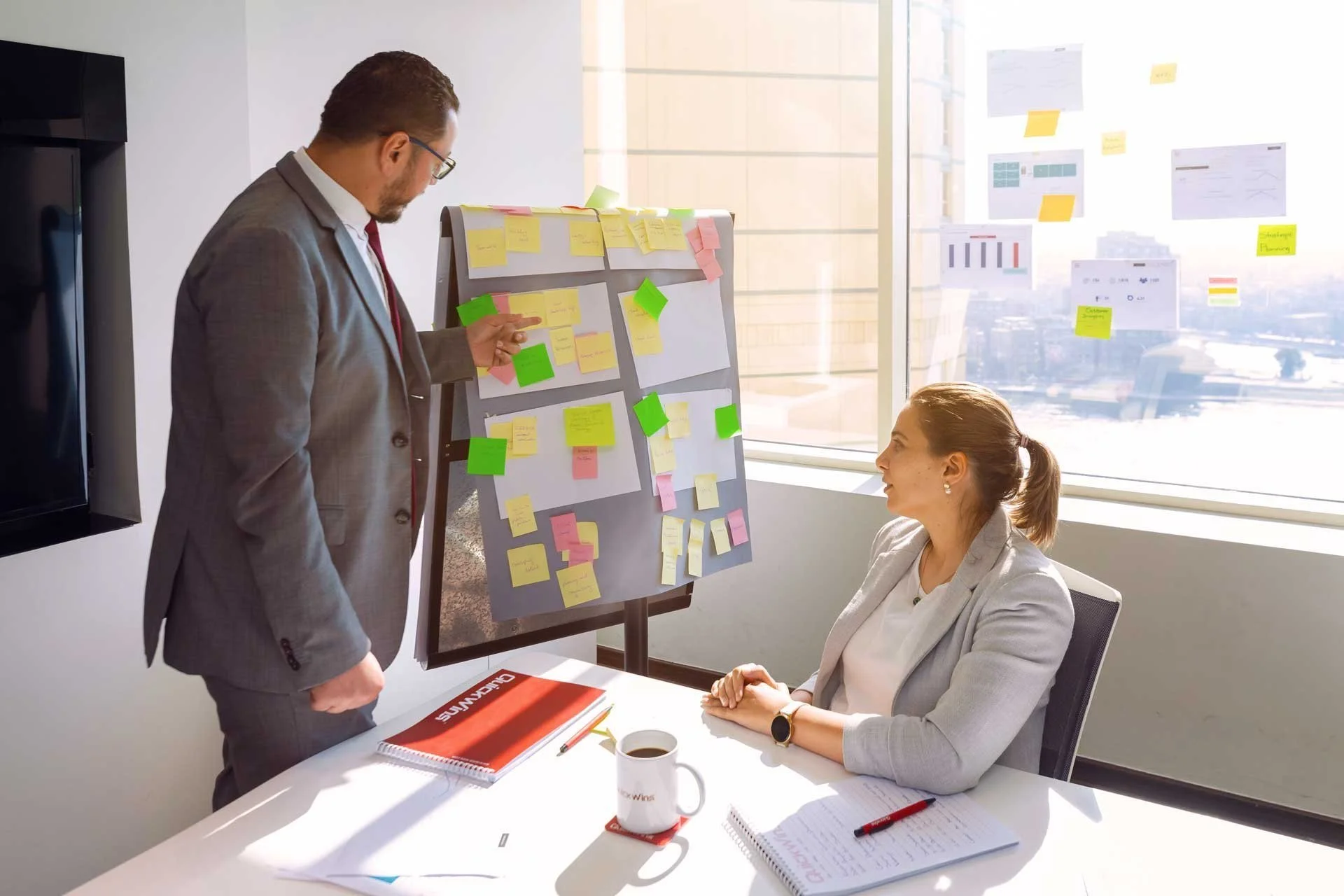 A man in a suit points to sticky notes on a planning board while a woman seated at the table listens.