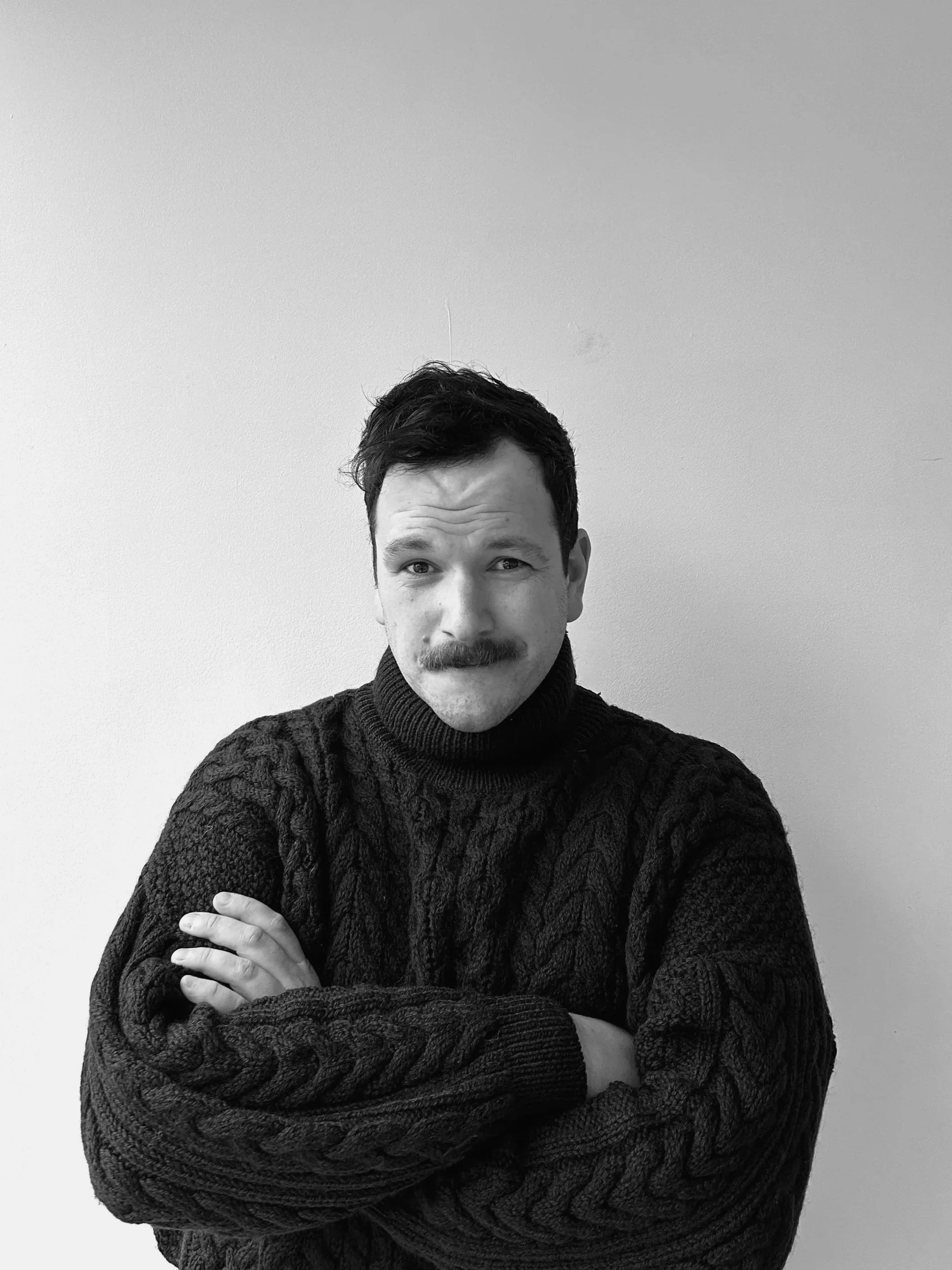 A man with dark hair, mustache, wearing a black cable-knit sweater, standing with arms crossed against a plain white wall.