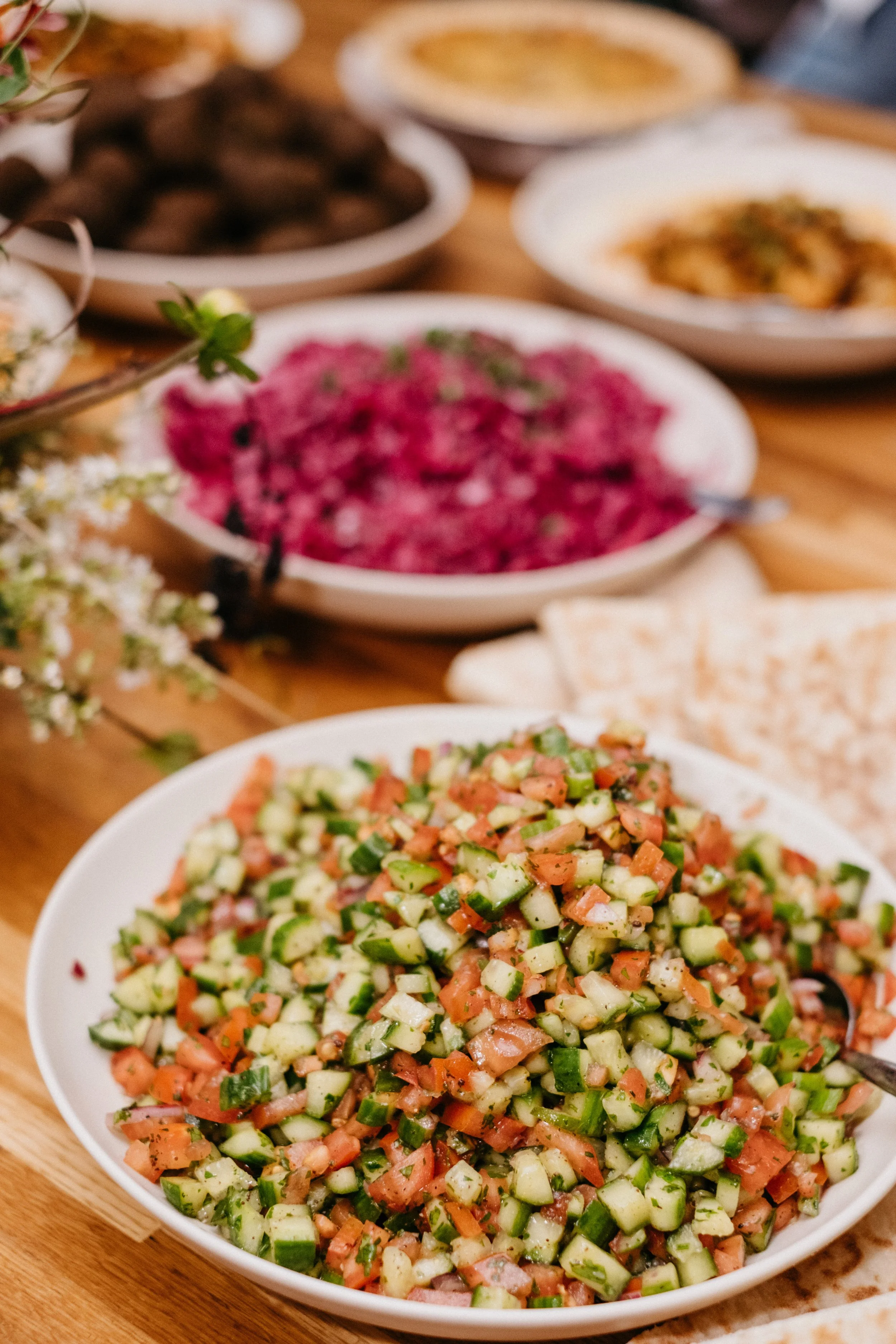 A bowl of chopped cucumber, tomato, onion, and herbs, garnished with black pepper, on a wooden table with other dishes in the background.
