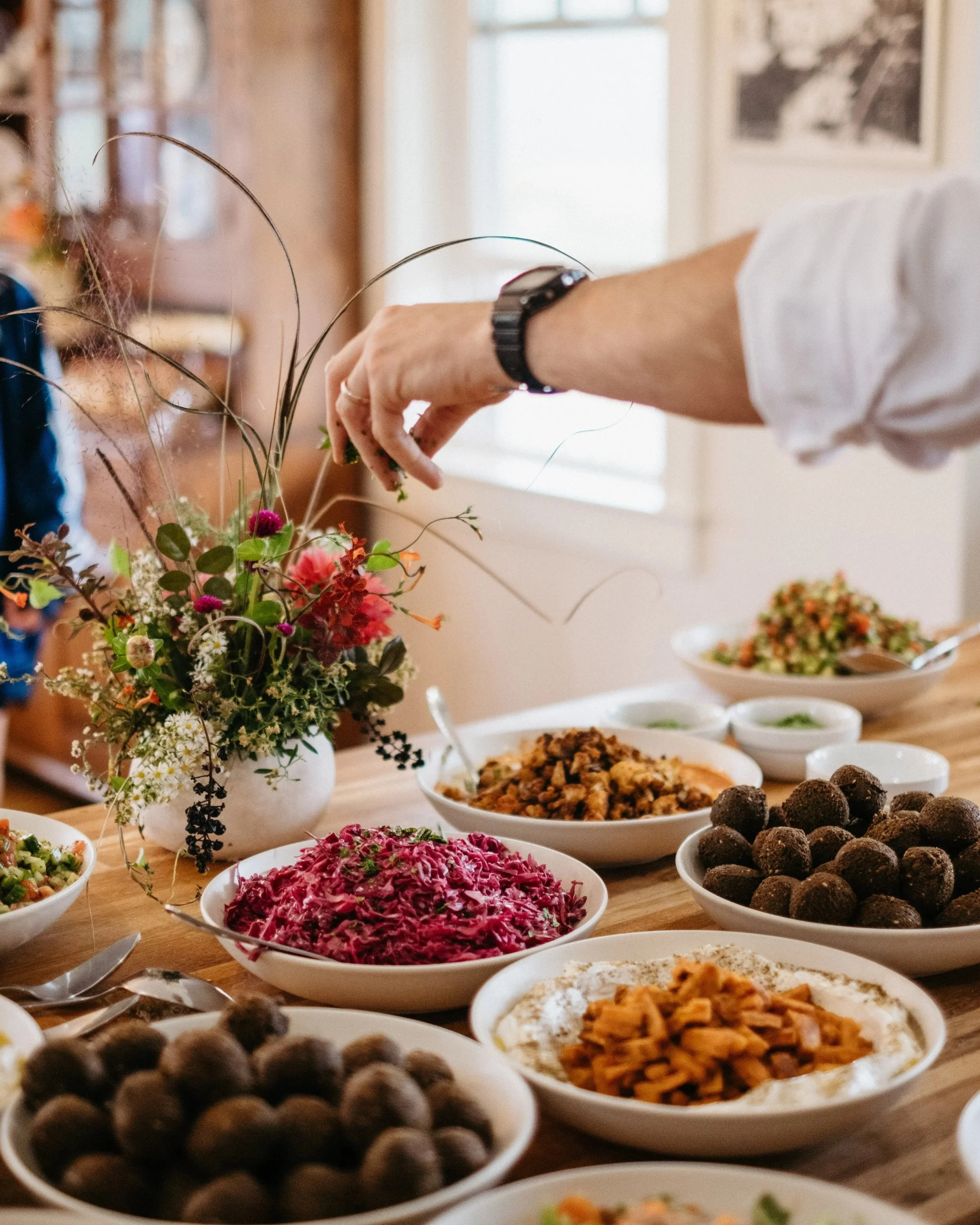 Person adding garnish to a flower arrangement on a dining table with various bowls of food including salad, beet salad, and appetizers.