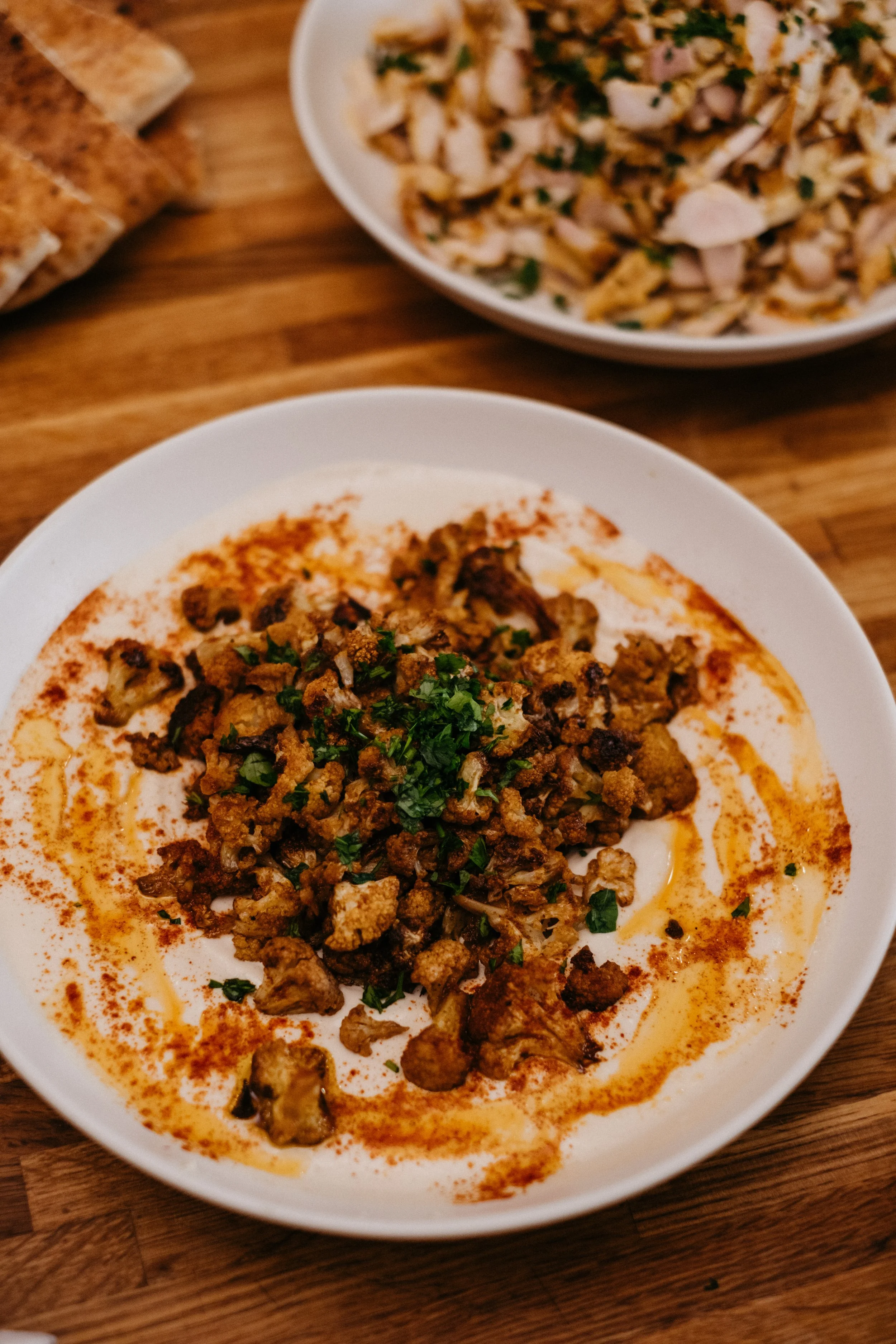 Close-up of a bowl of charred cauliflower with tahina with a bowl of shwarma in the background