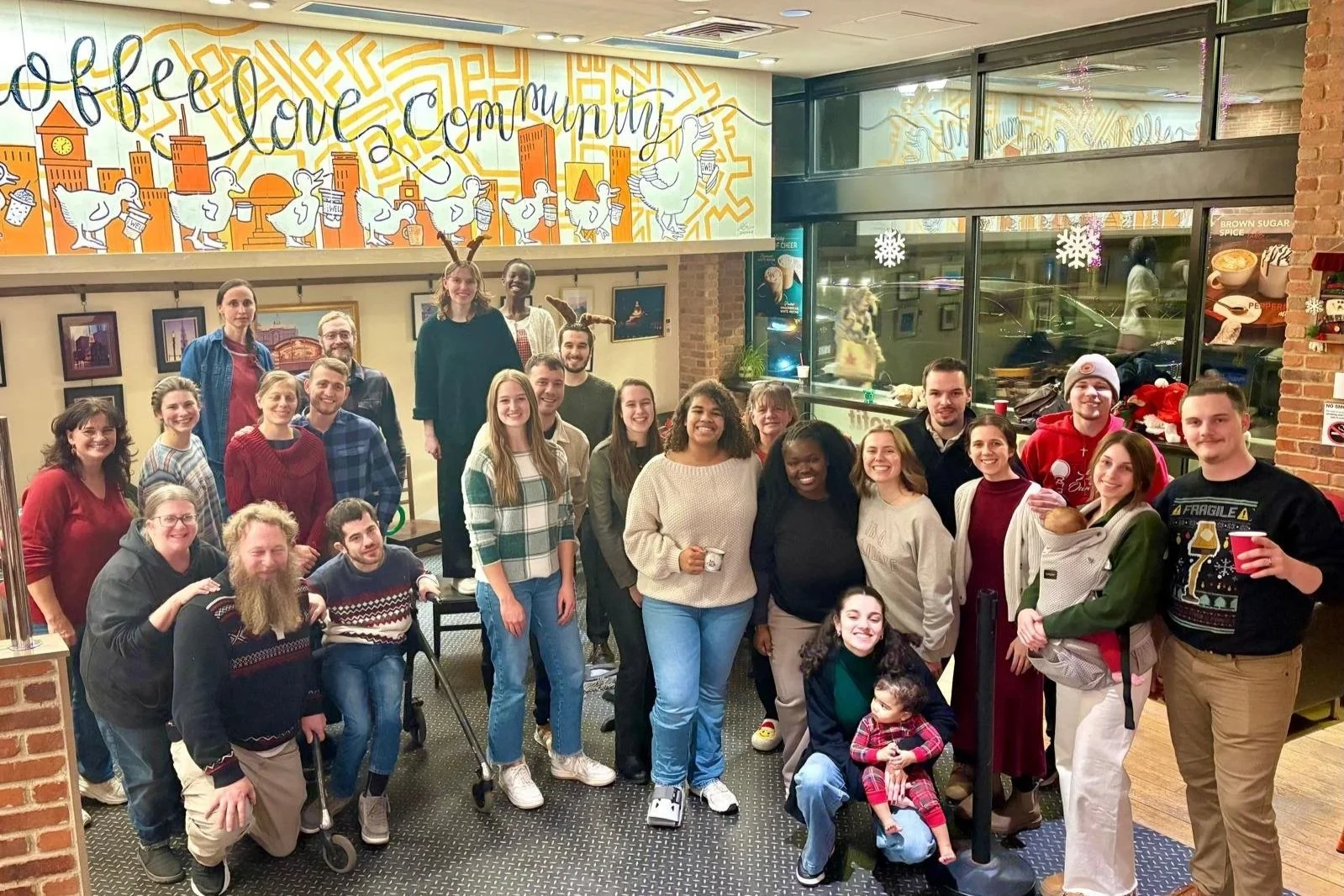 Group of people celebrating Christmas at The Well Coffee House, some wearing reindeer antlers and holiday sweaters, with festive decorations and artwork in the background.
