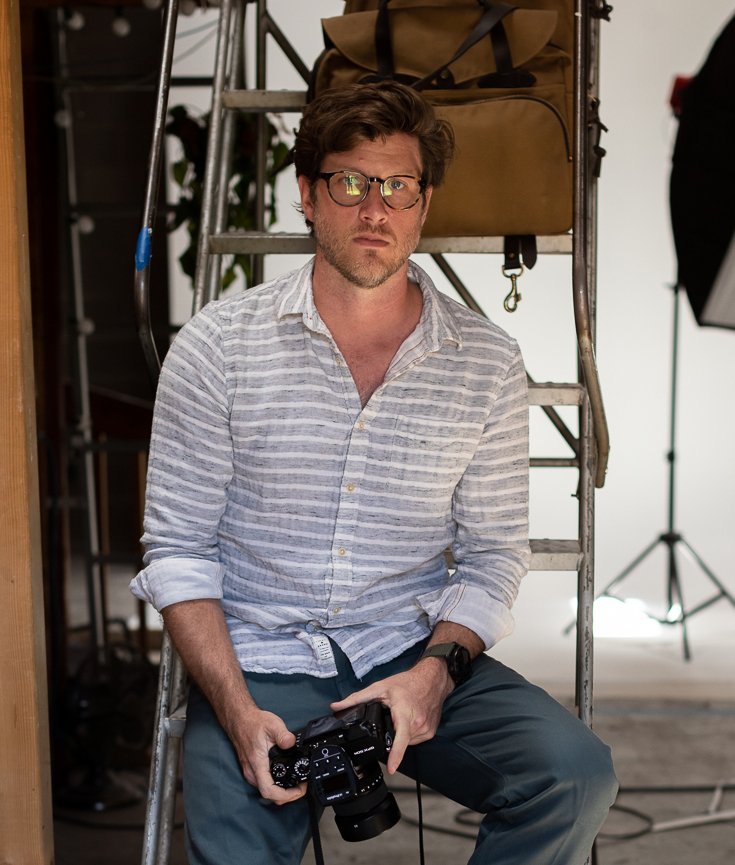 A man with glasses and a striped shirt sitting on a ladder holding a camera, with photography equipment in the background.