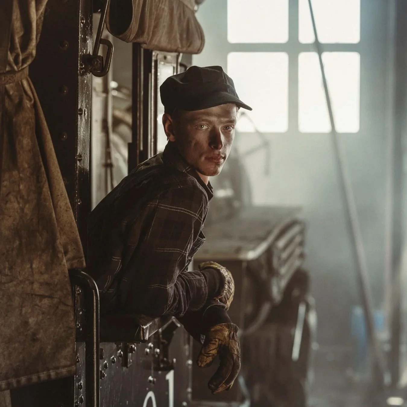 A young man with a dusty face and clothes, wearing a dark cap, leaning out of a train window in a dimly lit, dusty workshop or train depot with large windows in the background.