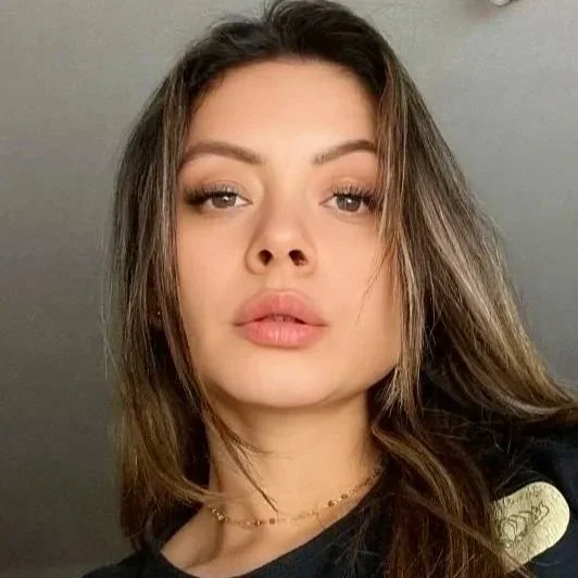 Close-up of a young woman with light brown hair, full lips, and subtle makeup, looking at the camera against a plain background.
