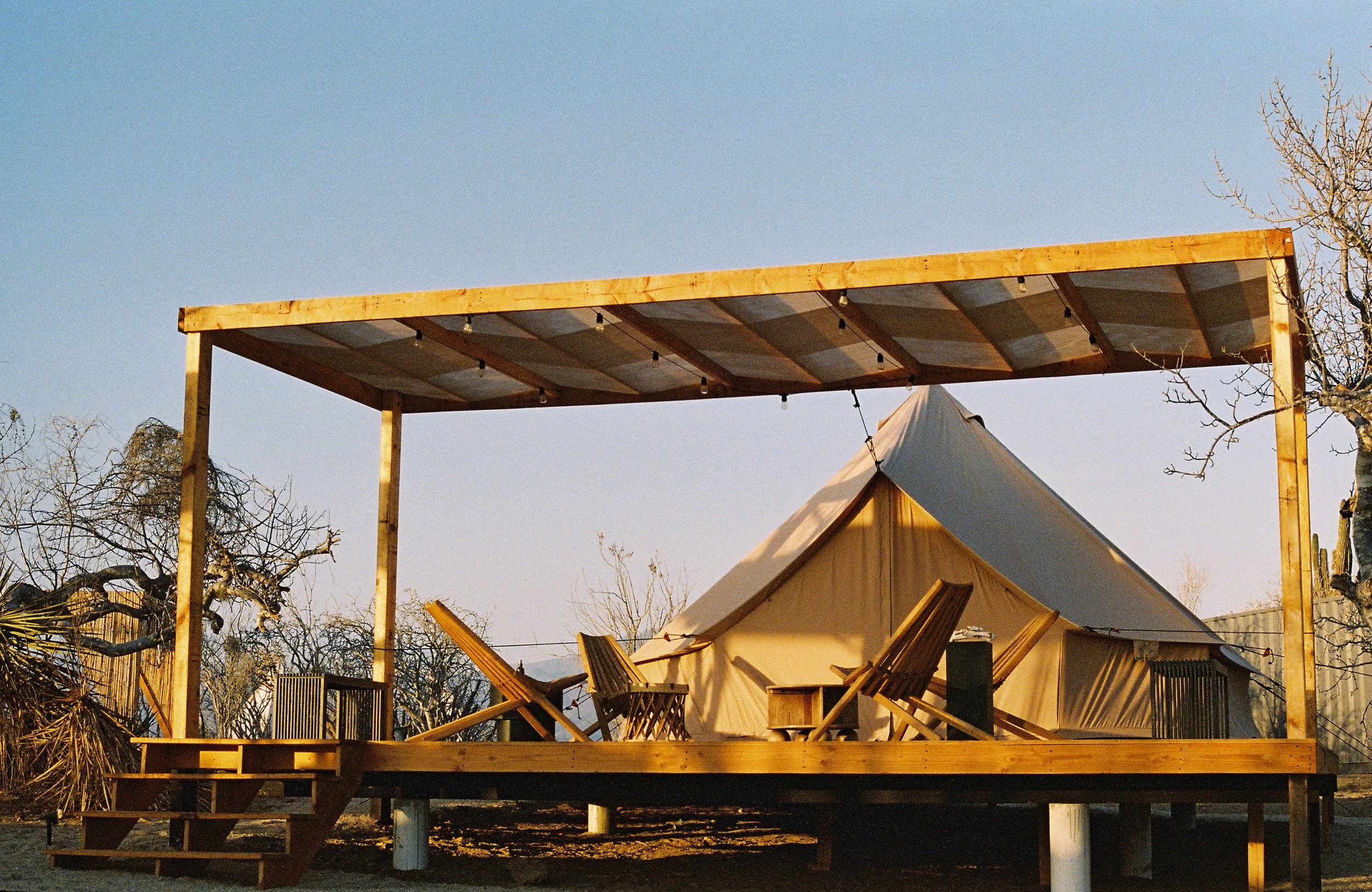 A desert landscape at sunset with a teepee tent on a wooden platform, surrounded by sparse bushes and cacti, with mountains in the distance and a pastel-colored sky.