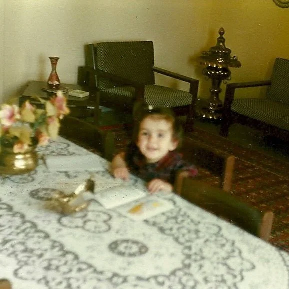 A young girl sitting at a dining table with a white lace tablecloth, a floral centerpiece, and small figurines. The room has vintage-style chairs and a rug on the floor.