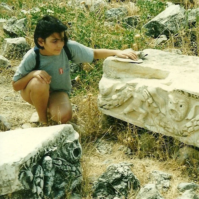 A young girl kneels on the ground outdoors, examining and touching ancient carved stone blocks with animal and human figures, surrounded by dry grass and scattered rocks.