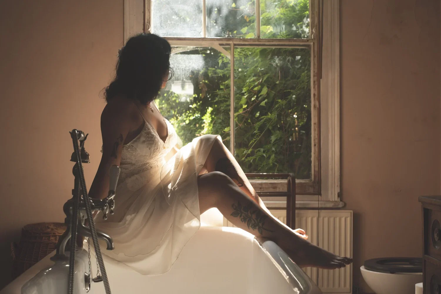 A woman in a white dress sitting on the edge of a bathtub in a bathroom, looking out a window with green foliage outside, with sunlight illuminating her and the scene.