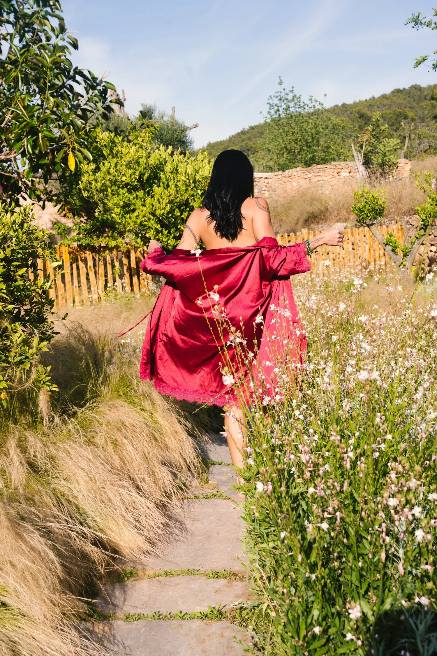 A woman with black hair, wearing a red dress, is walking along a garden path surrounded by wildflowers and greenery.