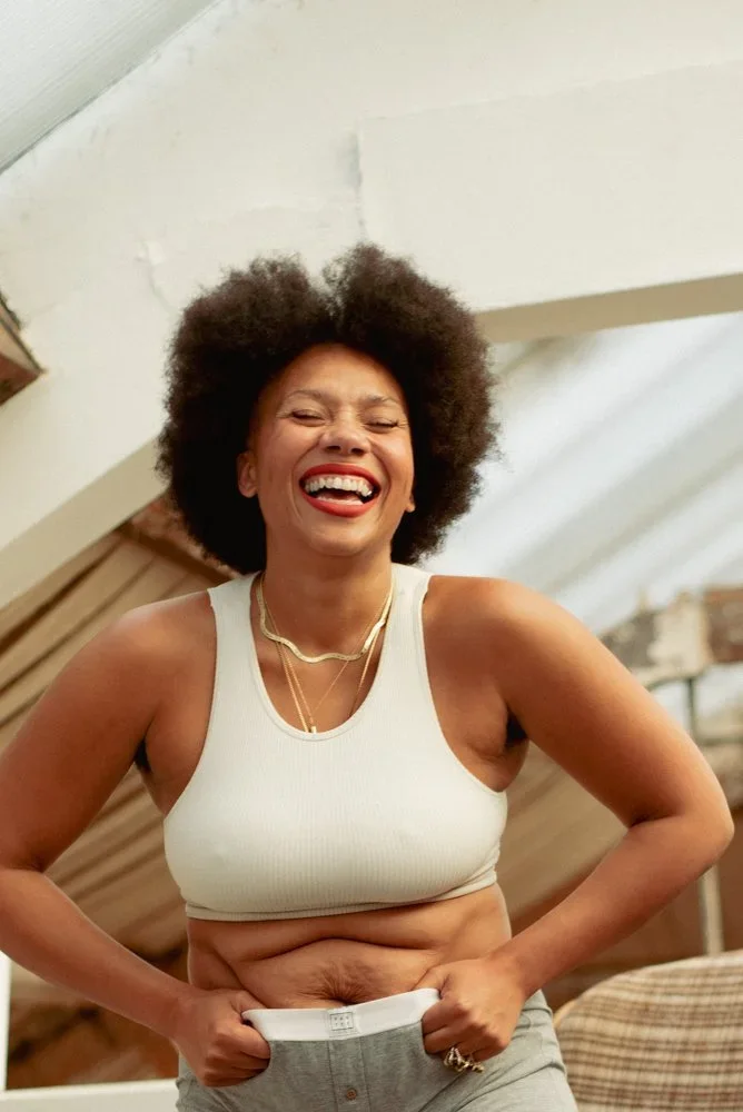 A woman with curly hair laughing, wearing a white crop top and gray pants, indoors with a light-colored ceiling.