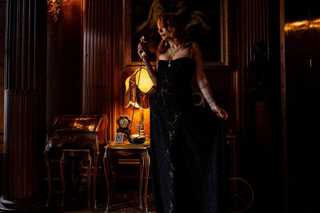 A woman with red hair stands in a regal room with a phone in one hand and dressed in an elegant black gown and golden jewellery