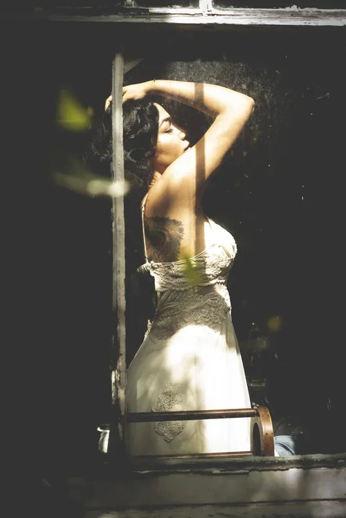 A woman with dark hair and a tattoo on her arm, wearing a white dress, standing by a window with her eyes closed and her arm raised above her head.