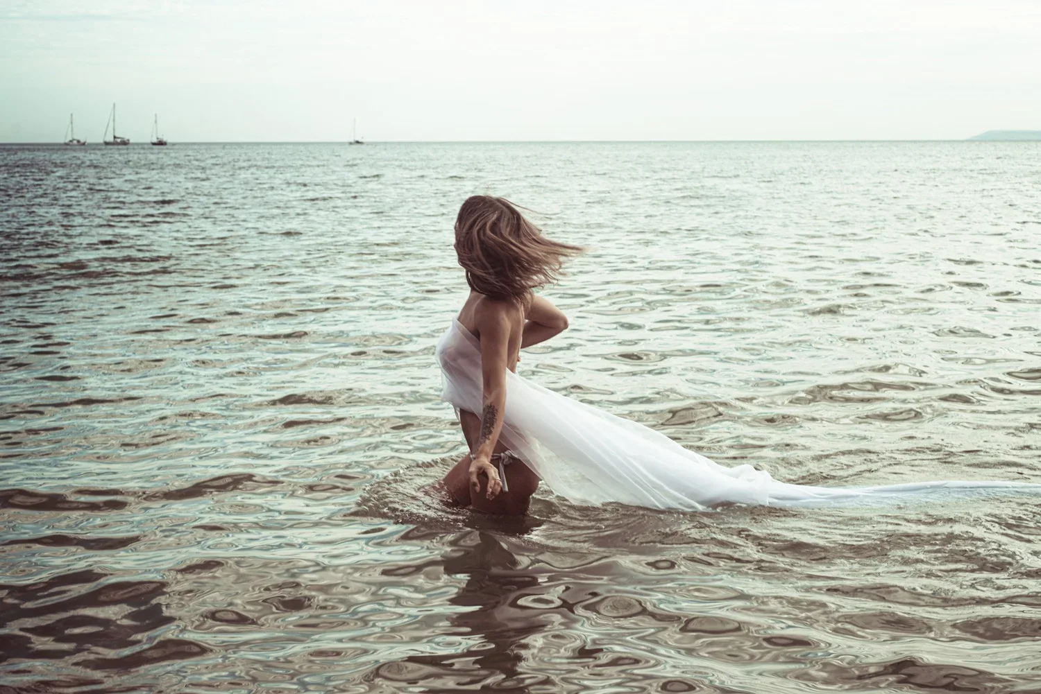 A woman in a white dress kneeling in shallow water at the beach, with her hair flowing and head turned to the side, against a backdrop of a cloudy sky and sailboats in the distance.