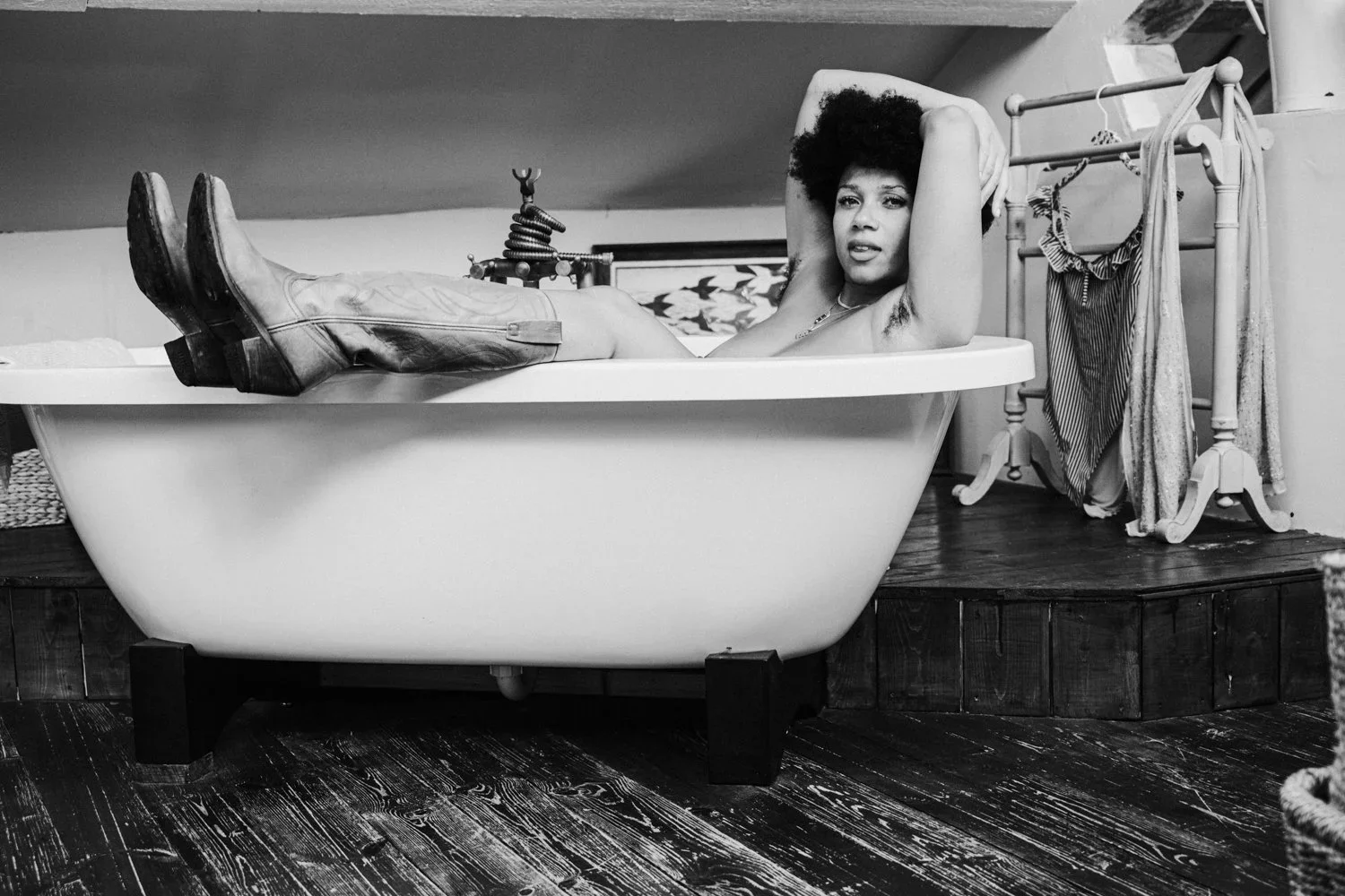 A woman with curly hair relaxing in a vintage bathtub, wearing cowboy boots. The bathroom has wooden flooring and a clothing rack with hanging clothes in the background.