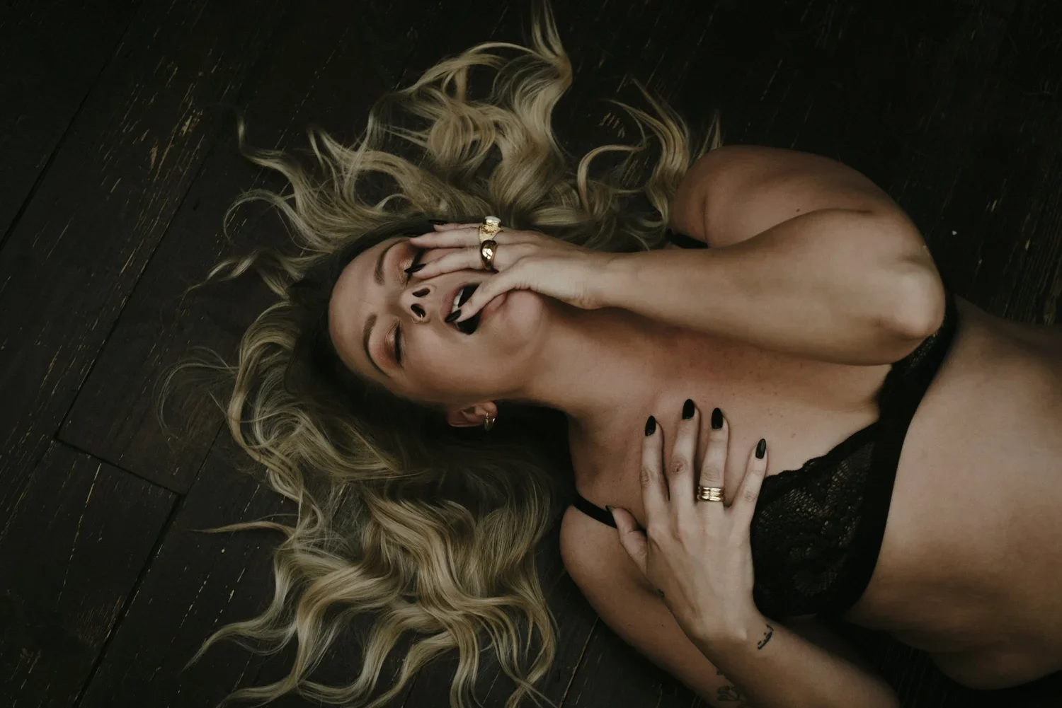 Woman lying on dark wooden floor with blonde wavy hair, black lingerie, and black nail polish, touching her face and chest, with eyes closed and an expression of pleasure or deep thought.