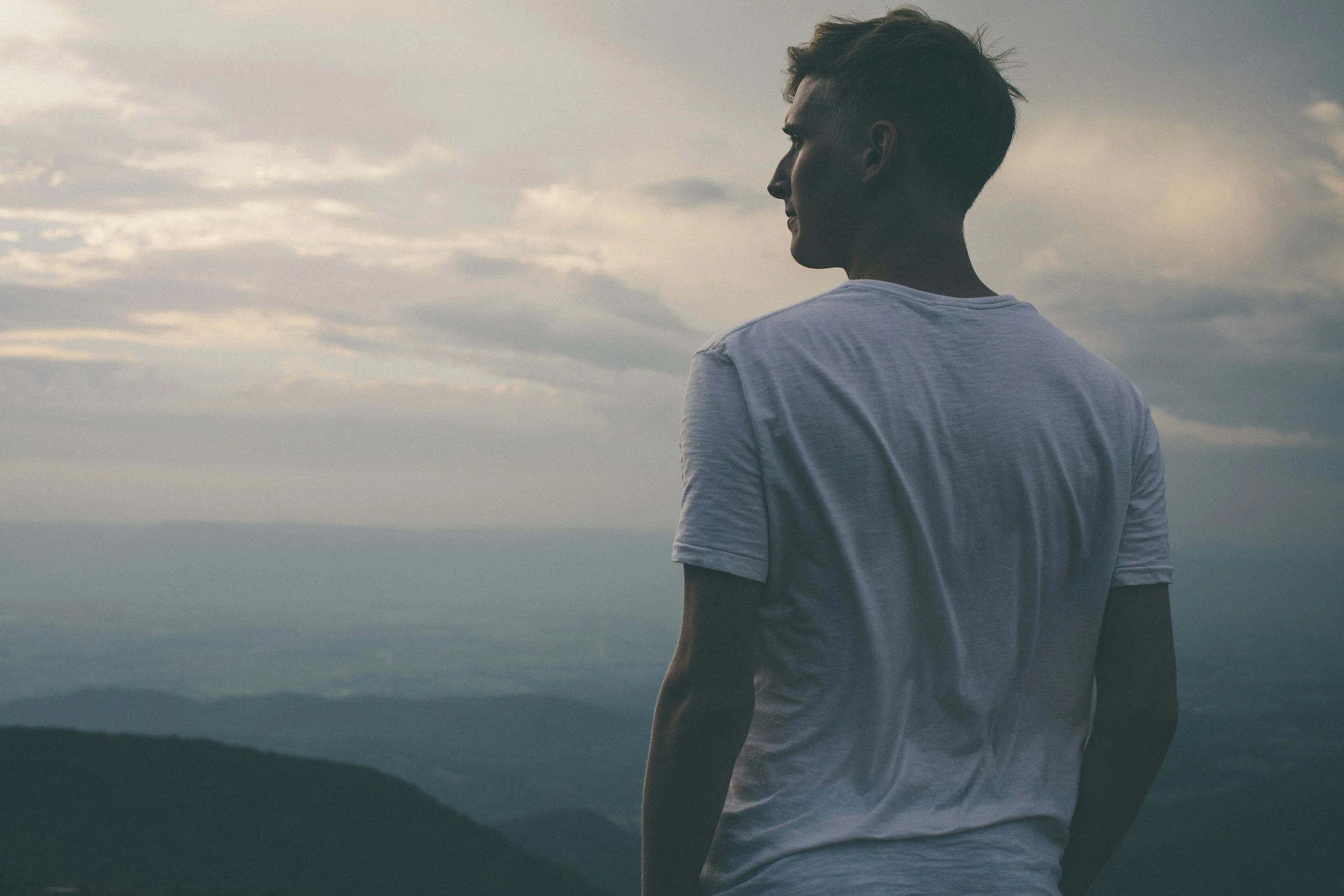 A young man with short hair wearing a white t-shirt, standing outdoors with a mountain landscape and cloudy sky in the background, looking away from the camera.