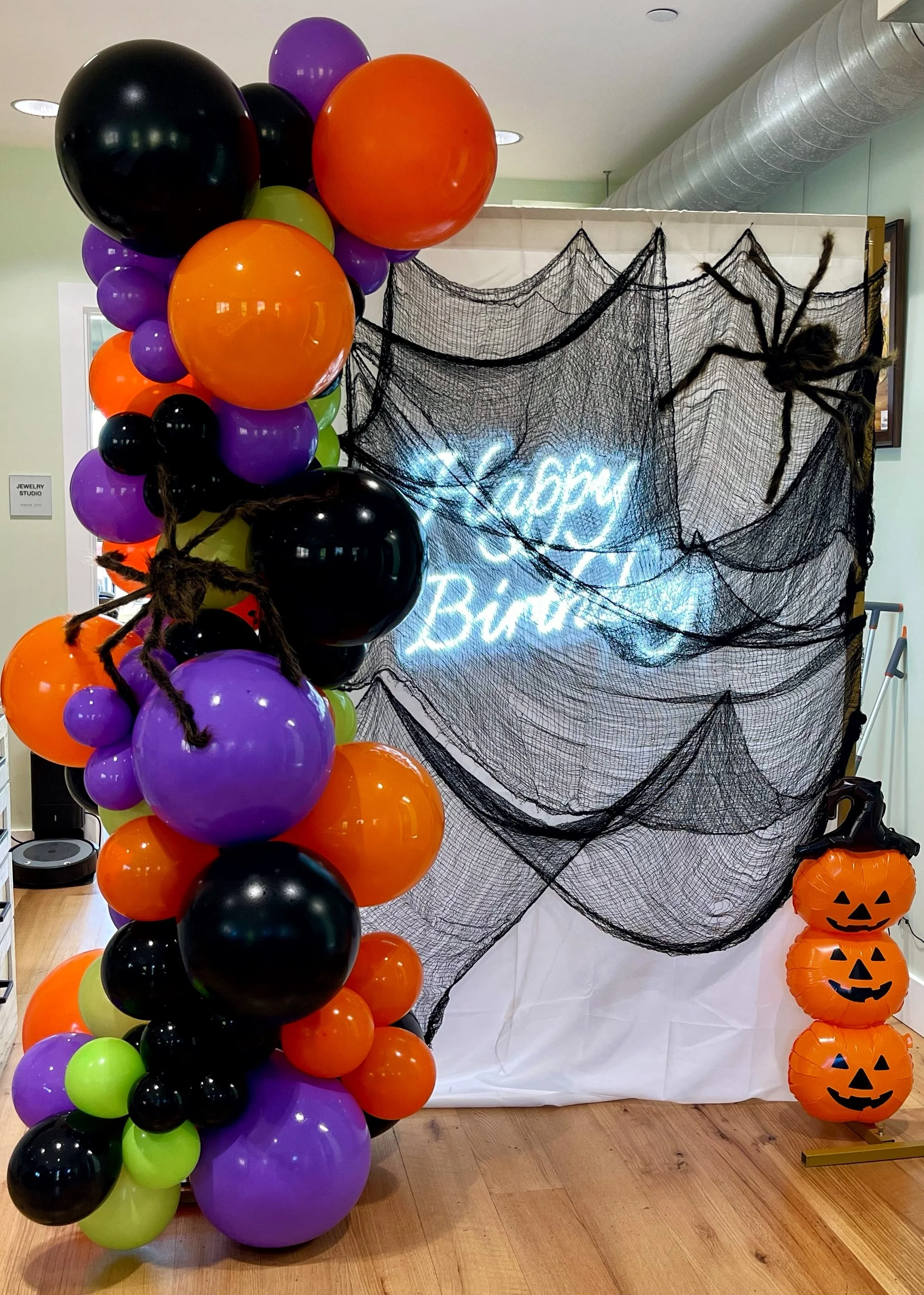 Halloween themed birthday party decoration with a balloon arch, fake spiders, a black net backdrop with an illuminated sign reading 'Happy Birthday,' and three stacked pumpkin-shaped balloons.