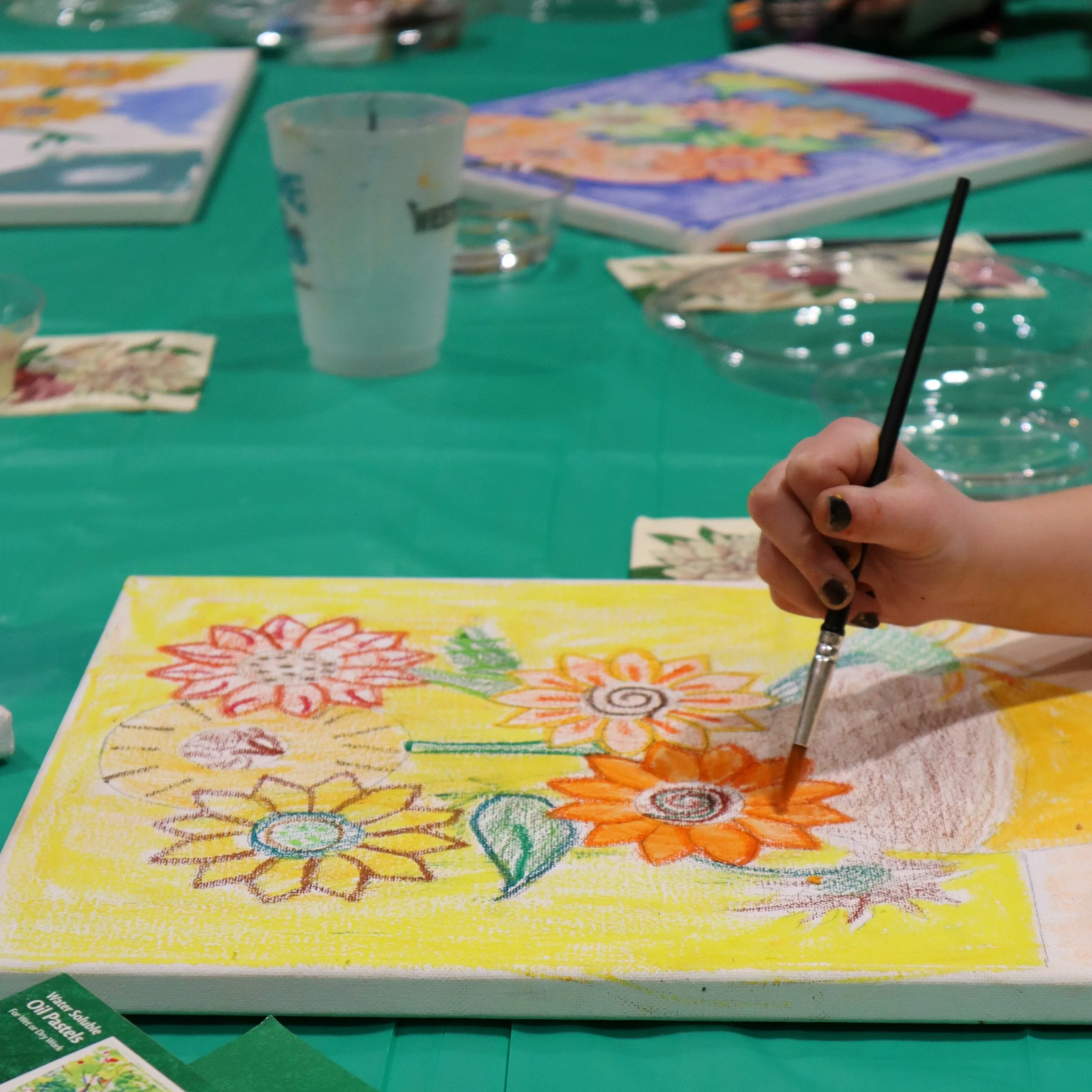 A person painting a colorful floral canvas with a paintbrush at a table, surrounded by art supplies and drink cups.