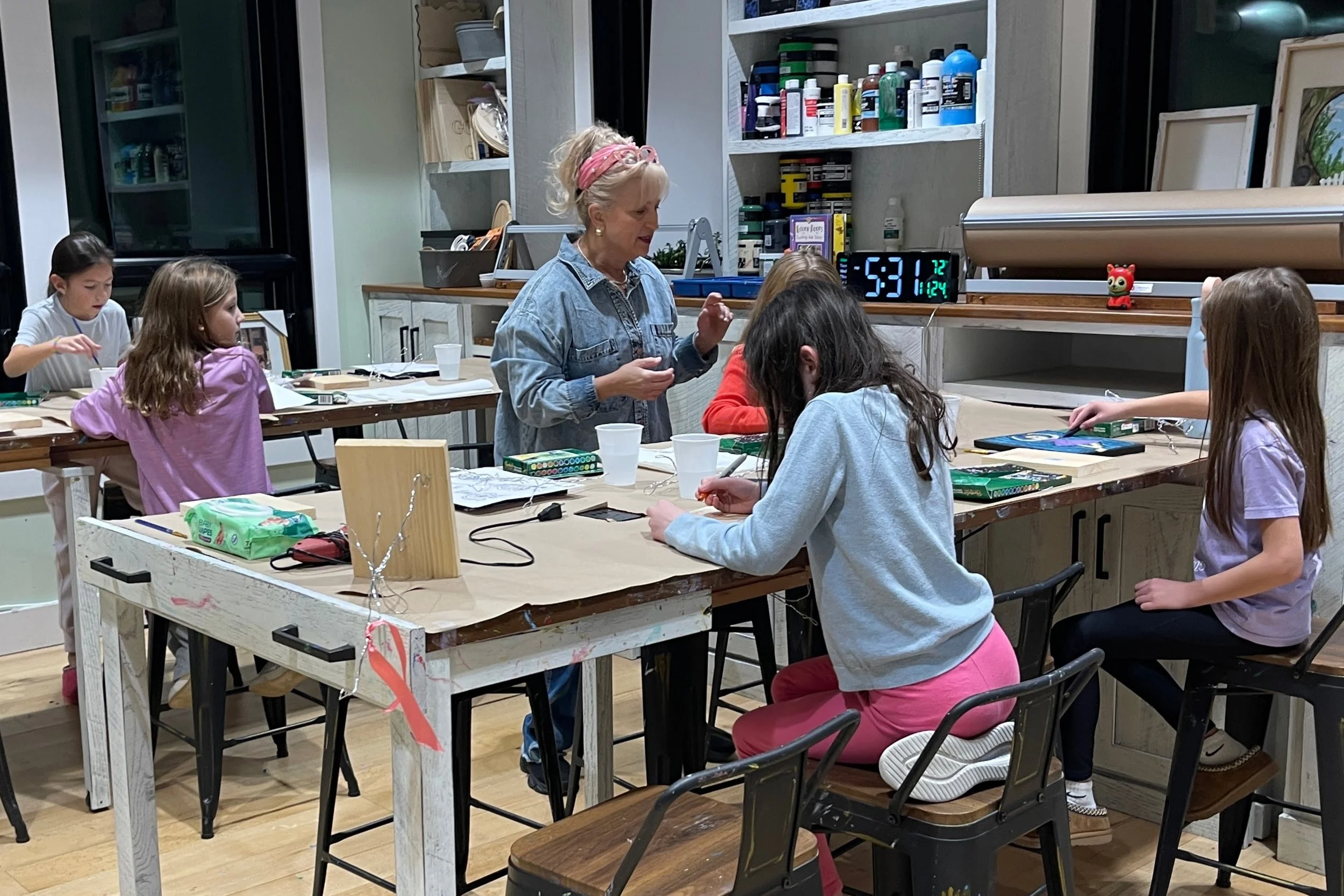 A group of children participating in an art class, sitting around a large wooden table with art supplies, and a woman guiding them inside a room with shelves of paint and art materials.