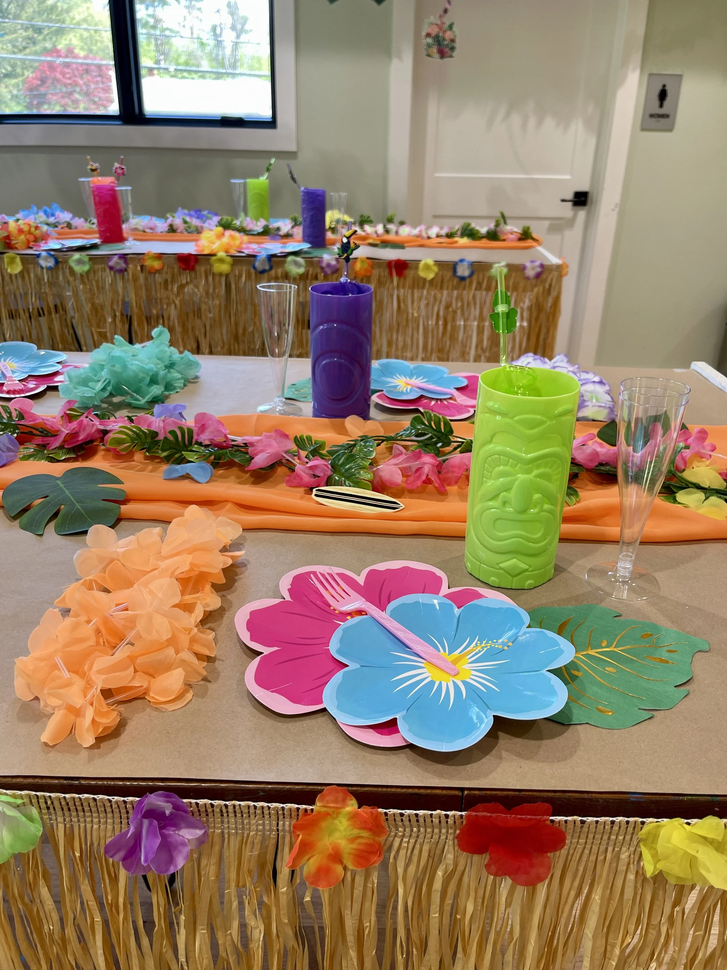 Colorful Hawaiian-themed table setup with paper flowers, tropical leaves, and drink dispensers in bright colors.