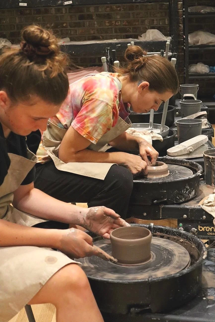 Two women are sitting at pottery wheels, shaping clay into pottery pieces. They are focused on their work, with pottery tools and supplies around them.