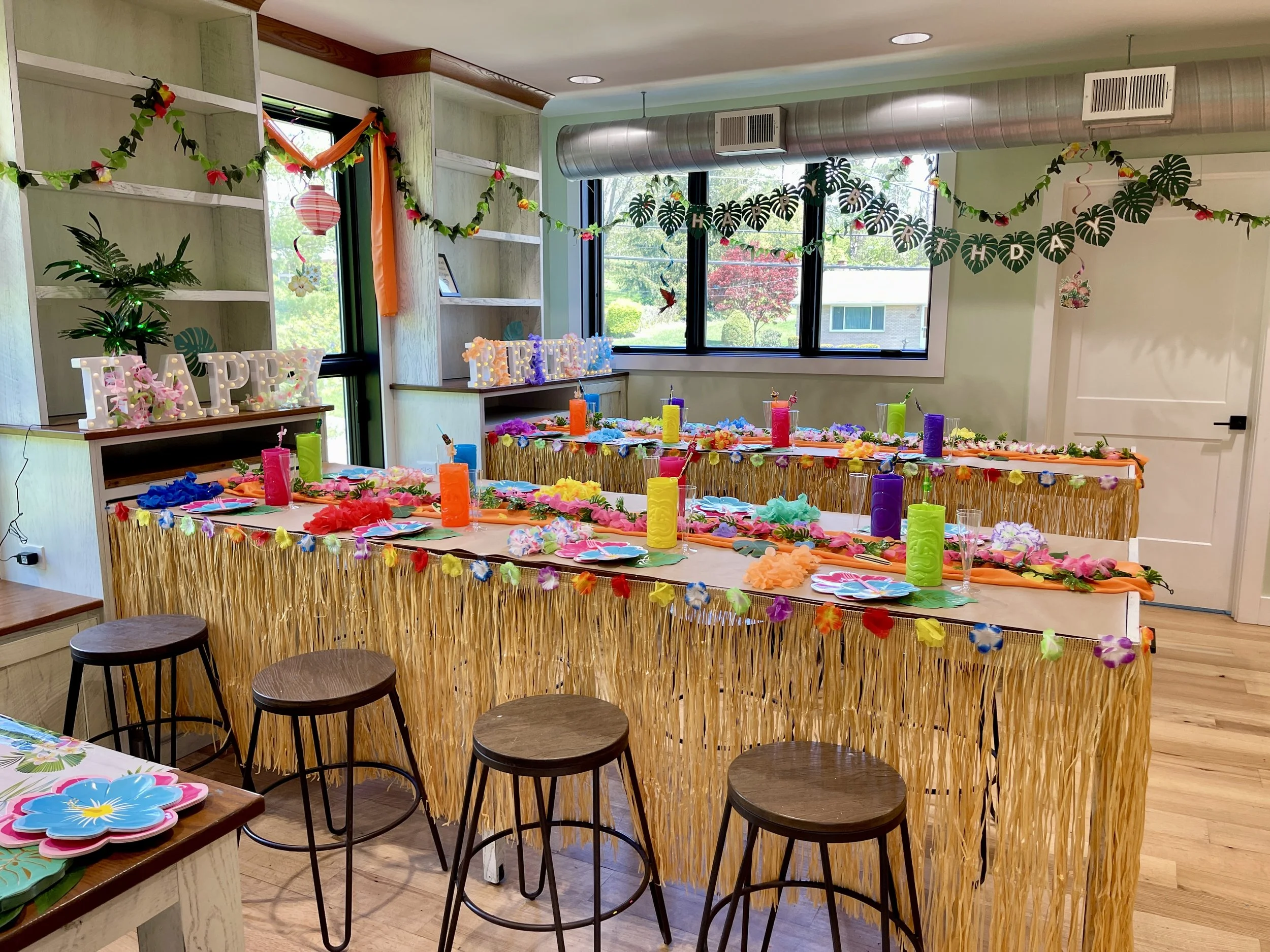 Colorful tropical-themed party setup featuring tables decorated with floral leis, tiki torches, and paper flower plates. The room has a