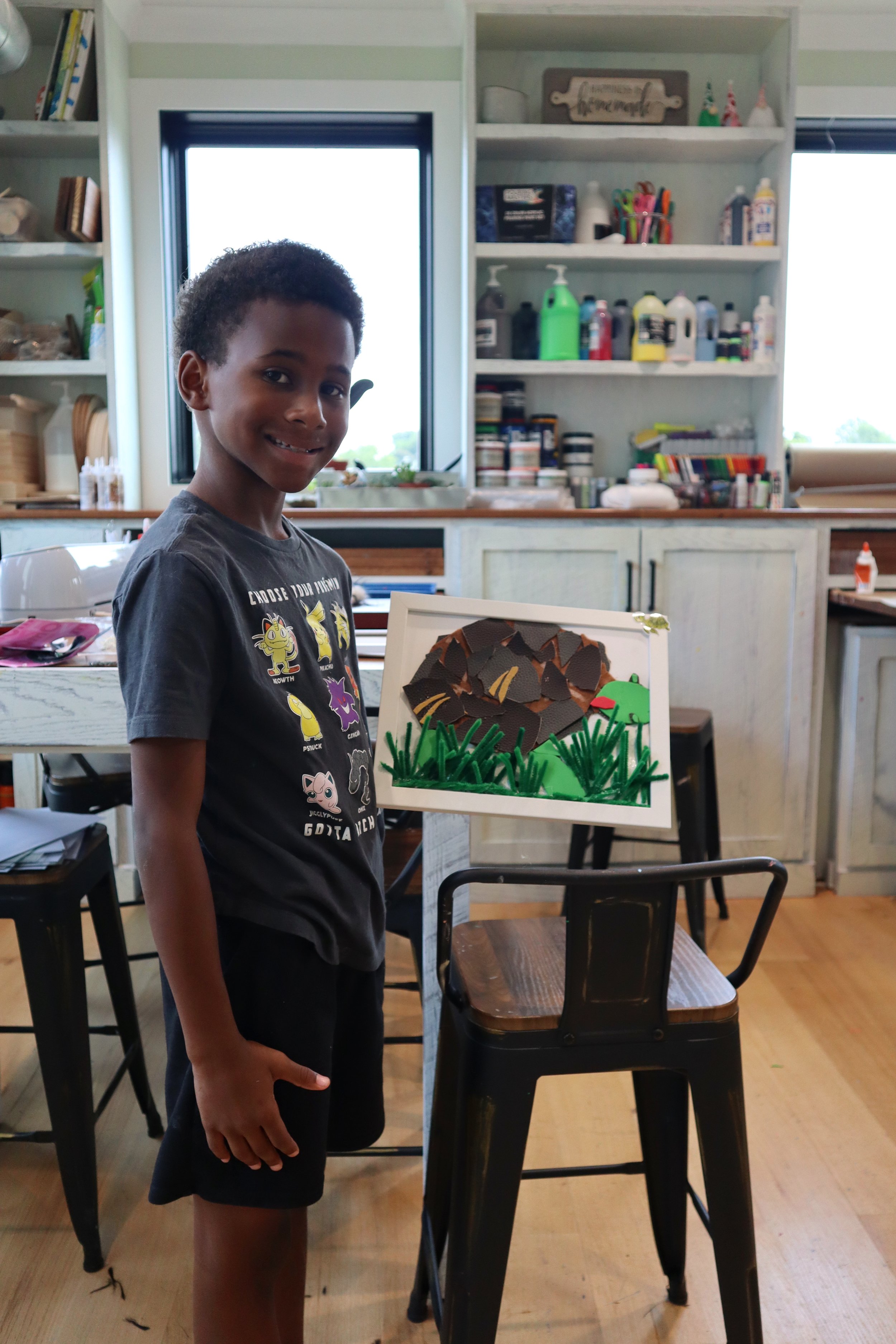 A boy standing indoors behind a small table, displaying a diorama of a forest scene with a bear made of paper and other craft materials. The background shows shelves with art supplies and windows.