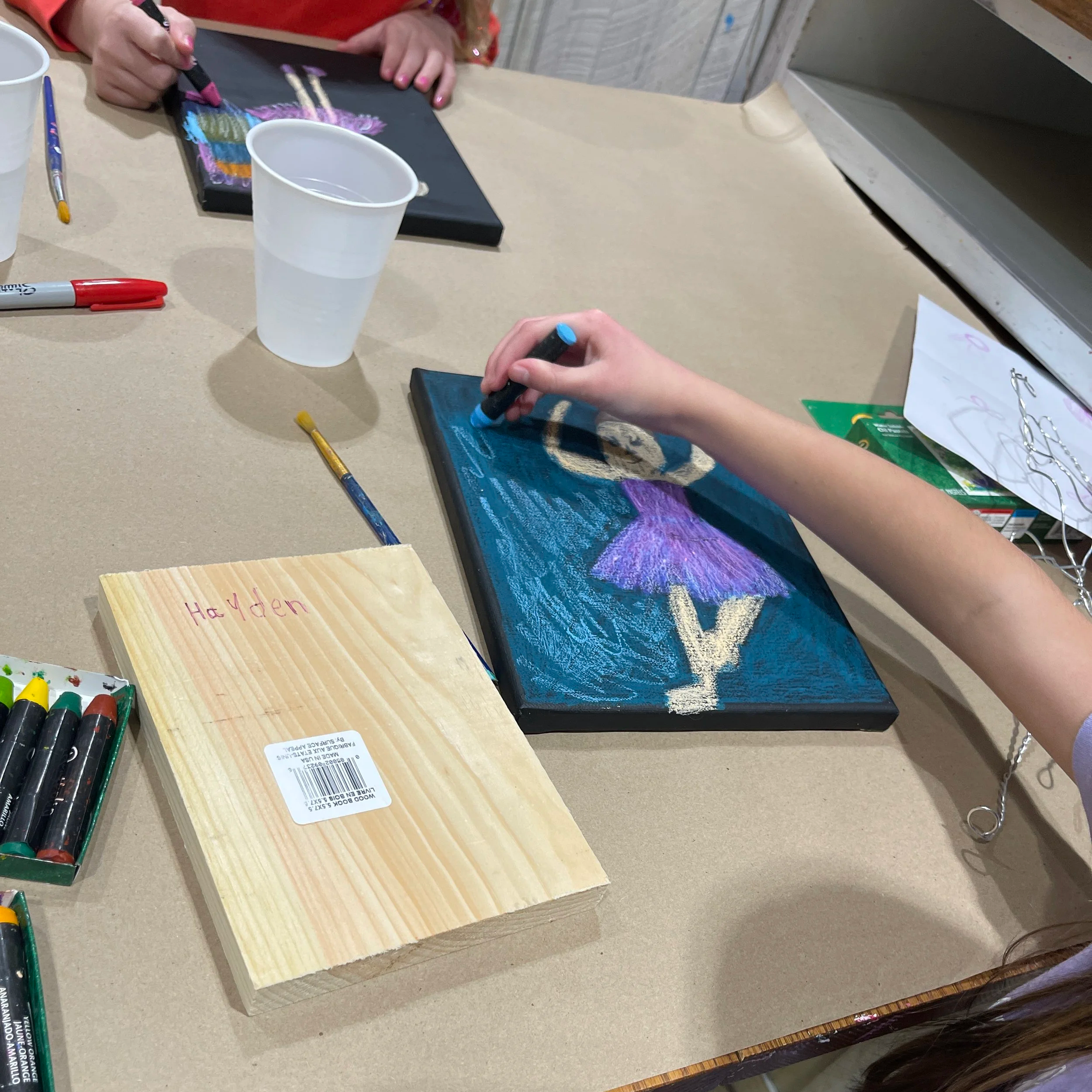 Child's hands drawing a picture of a girl in a purple dress with a crescent moon in a night sky on a small canvas with chalk, surrounded by art supplies like markers and a wooden block labeled 'Hayden'.