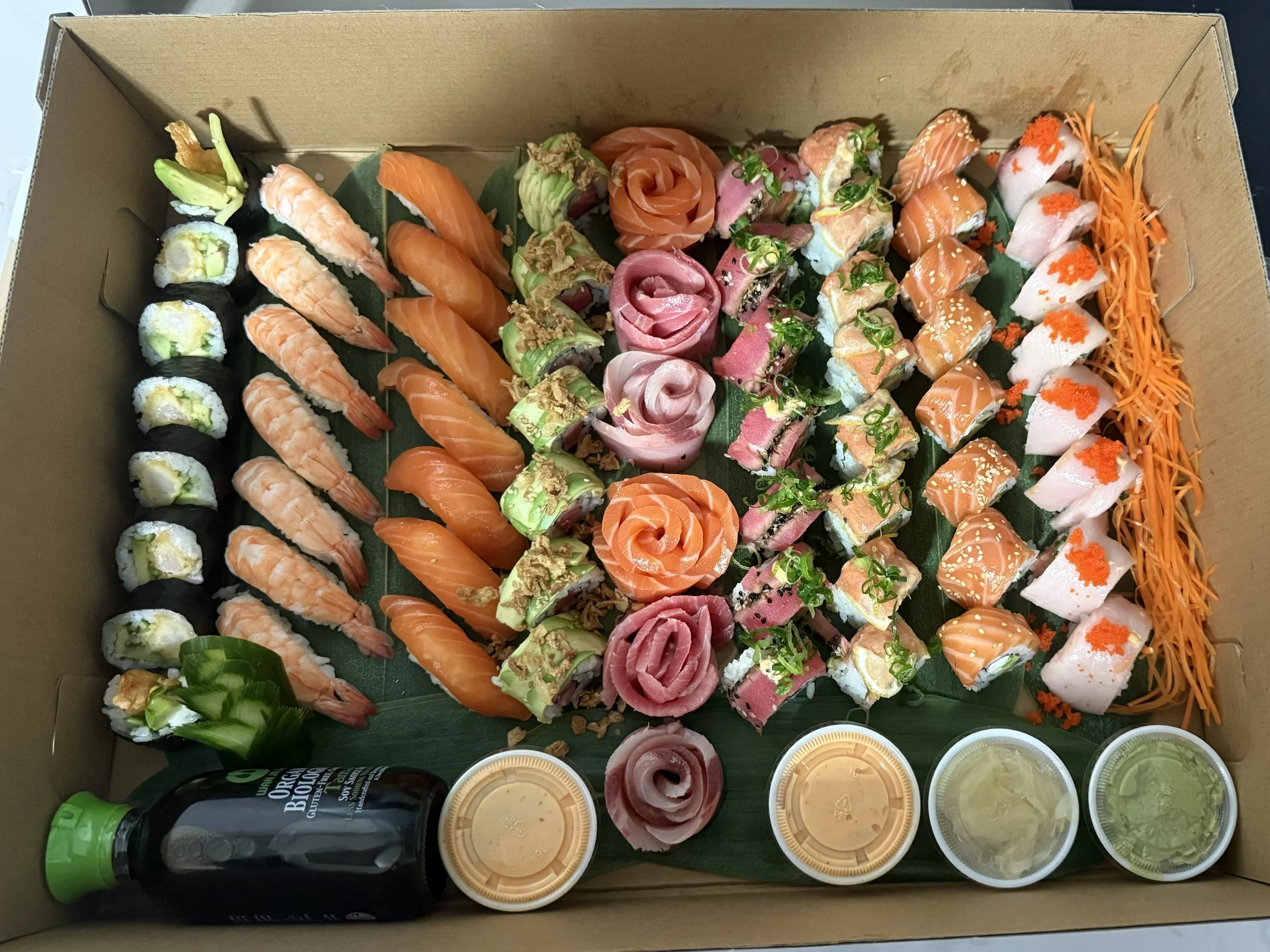 Assorted sushi rolls, sashimi, and Japanese appetizers in a cardboard box with small containers of dipping sauces and drinks.